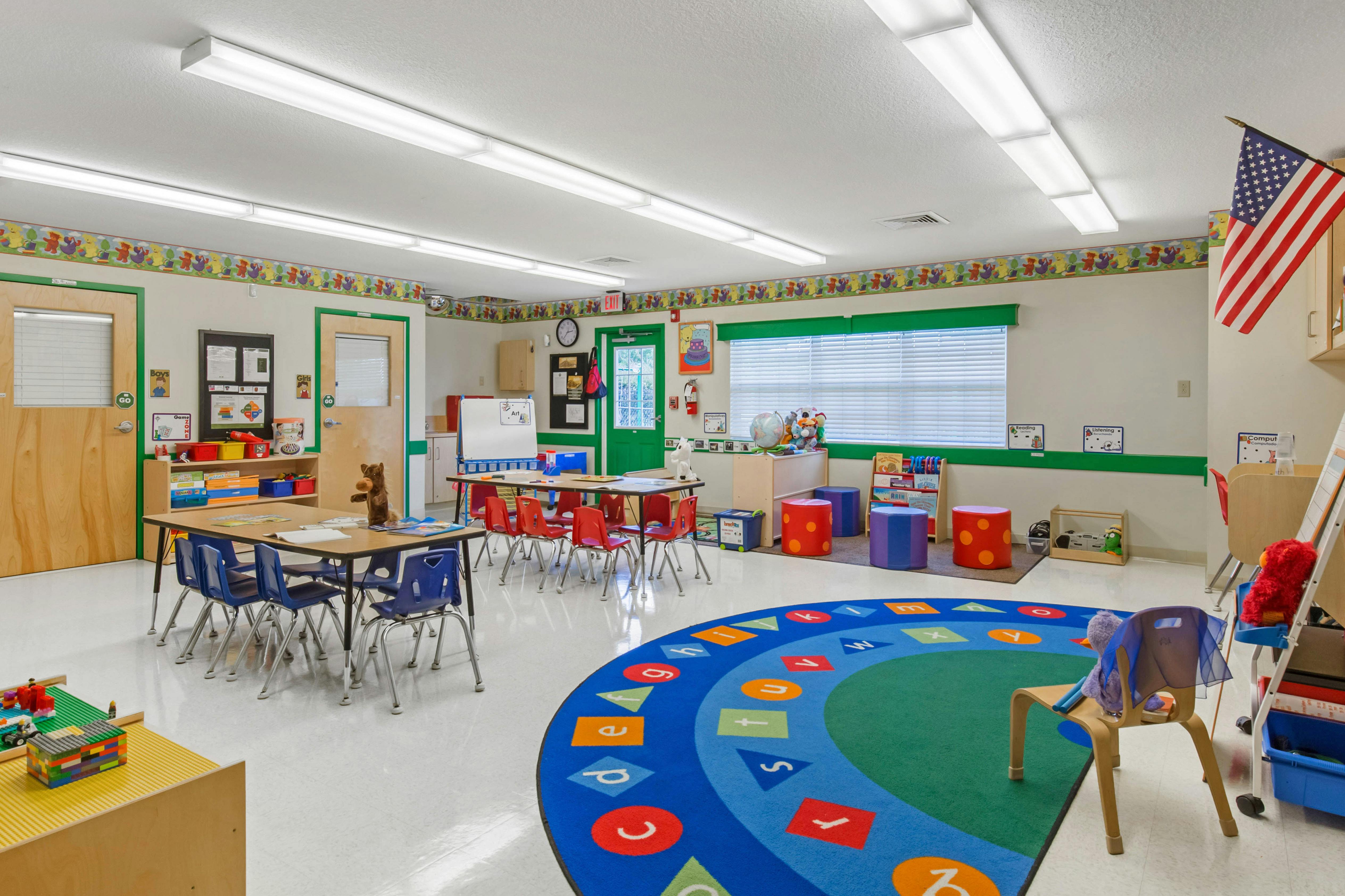 A colorful empty classroom with small tables and chairs, a play rug, and an American flag