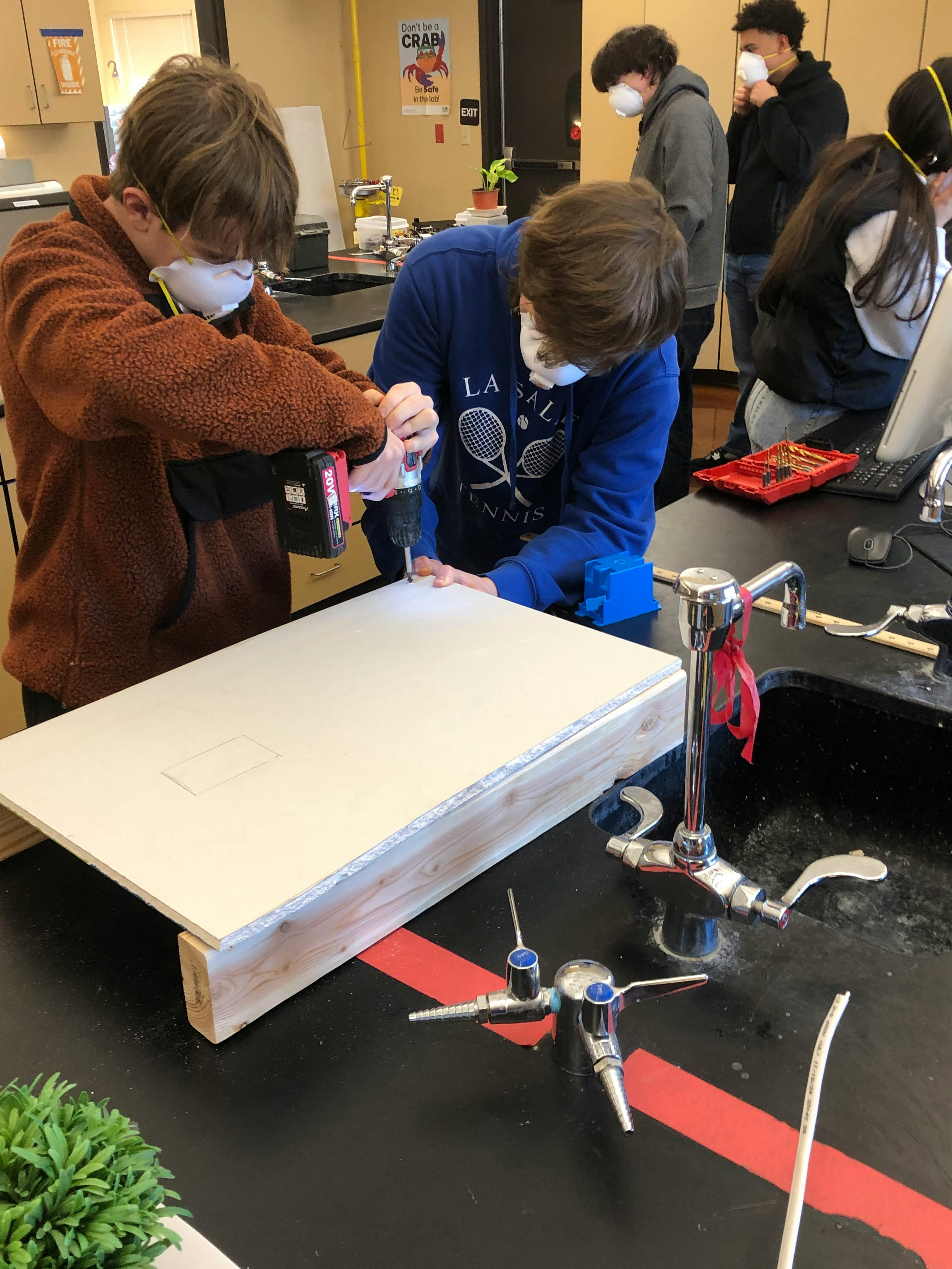 Two teens using a drill on a piece of wood in a classroom