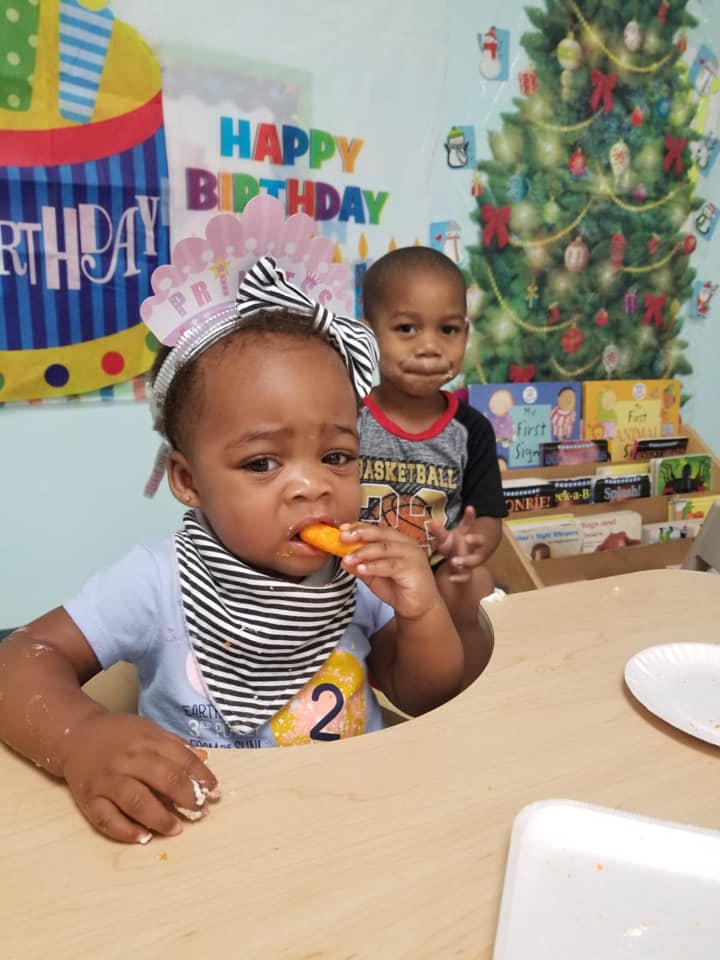 Child in a high chair eating a carrot stick, another child behind