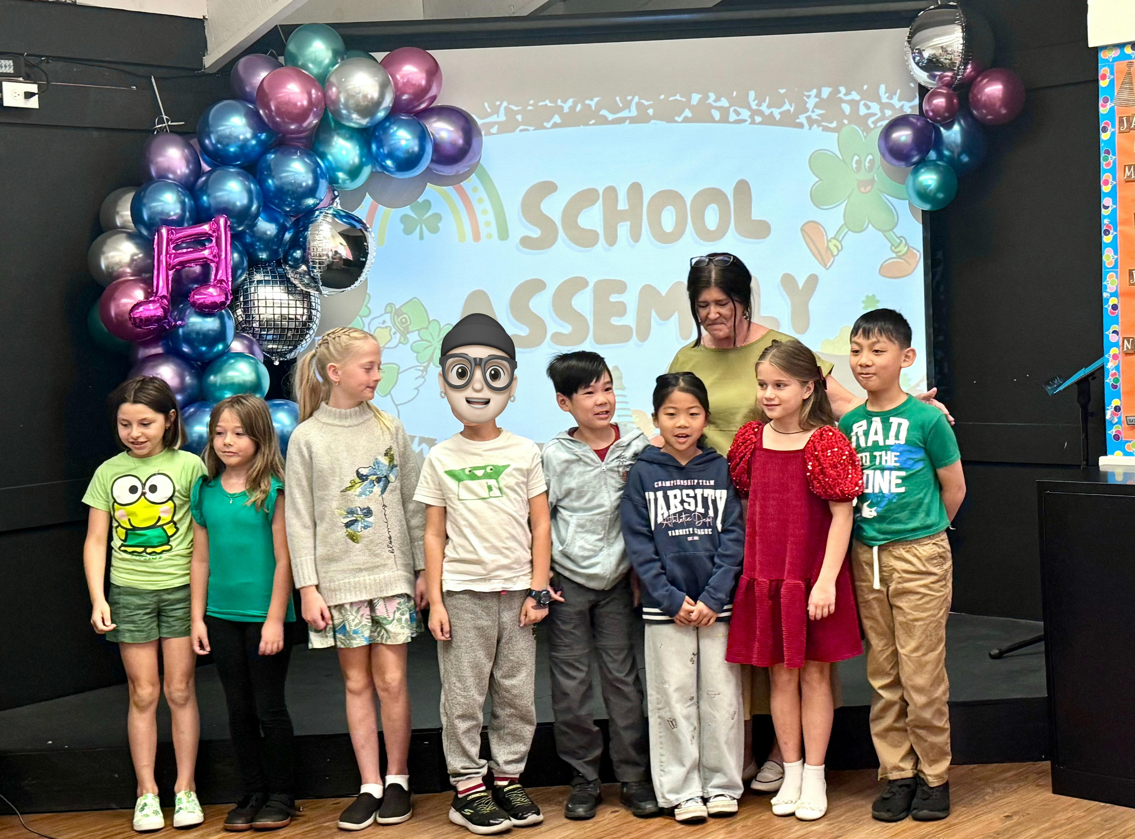 Teacher and six children posing in front of a screen that says "SCHOOL ASSEMBLY"