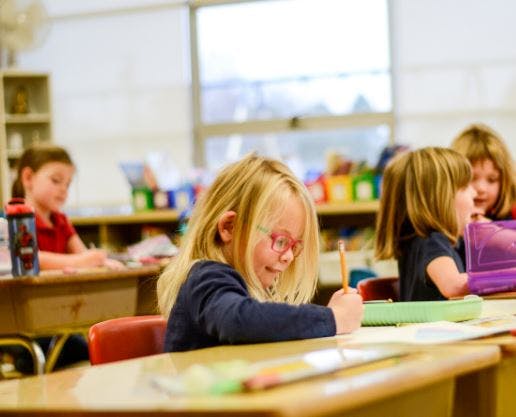 Child with glasses writing at a desk in a classroom
