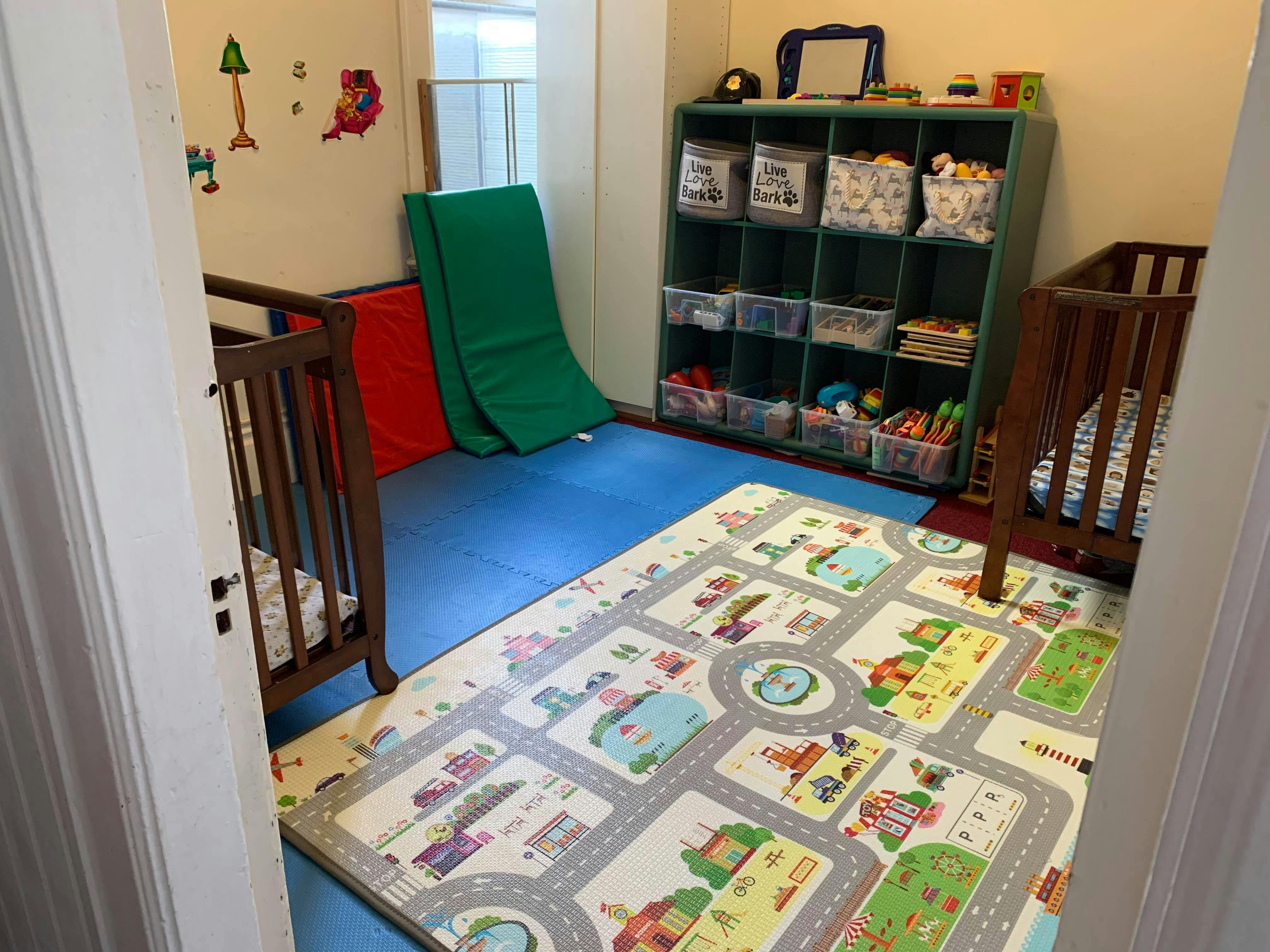 Playroom with two cribs, a toy shelf, and a playmat on the floor
