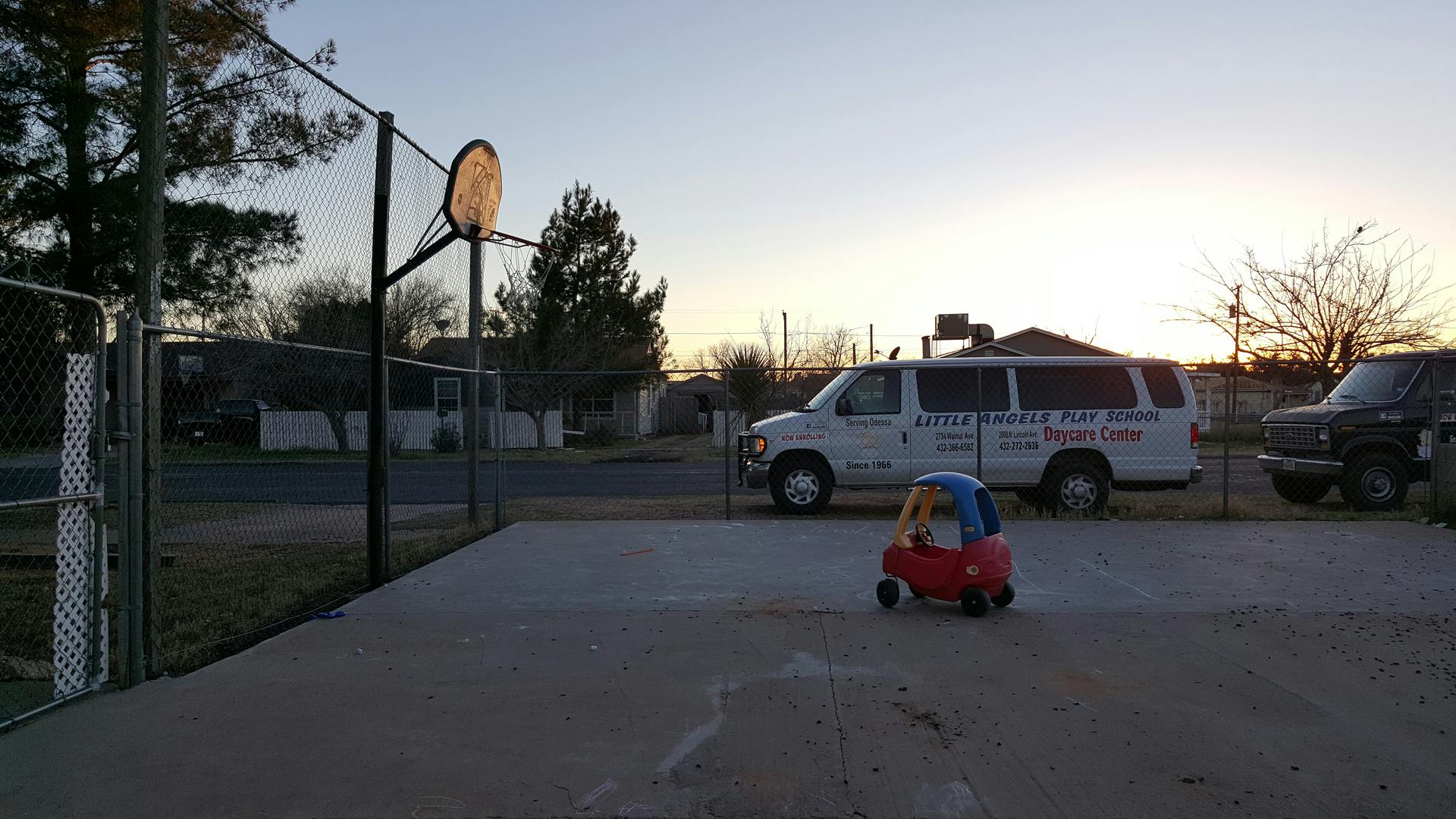 Toy car on a paved area with a basketball hoop and vehicles in the background
