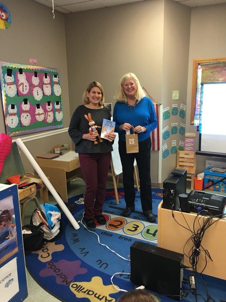 Two people standing in a classroom, one holding a book and toy, the other a small bag