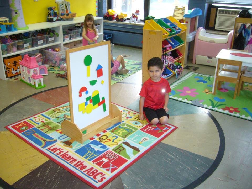Child kneeling on a rug in a classroom with another child standing behind an easel
