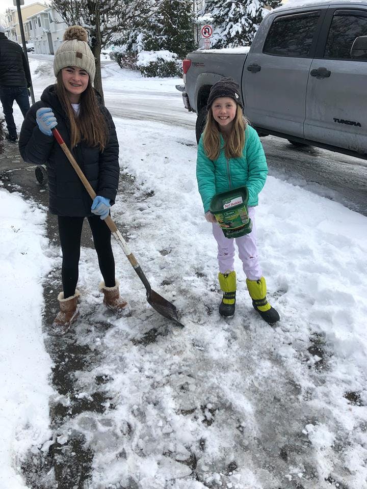 Two children on a snowy sidewalk, one holding a shovel and the other a bucket