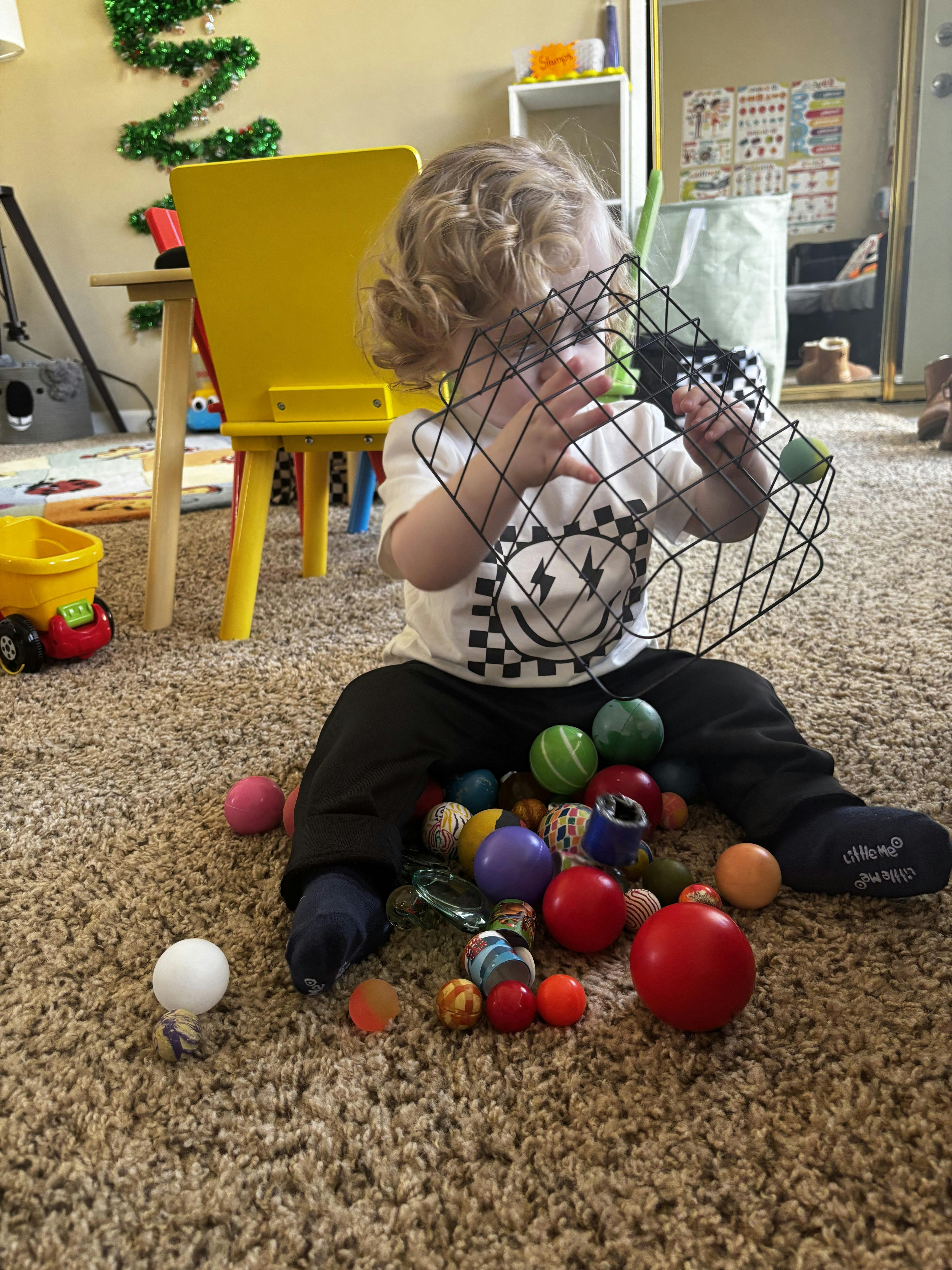 Child playing with a wire basket and colorful balls on a carpeted floor