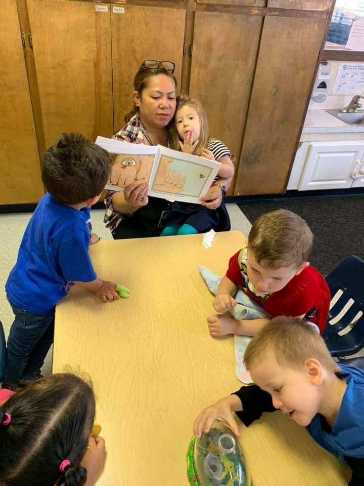 Person reading a book to a child, with other children at a table