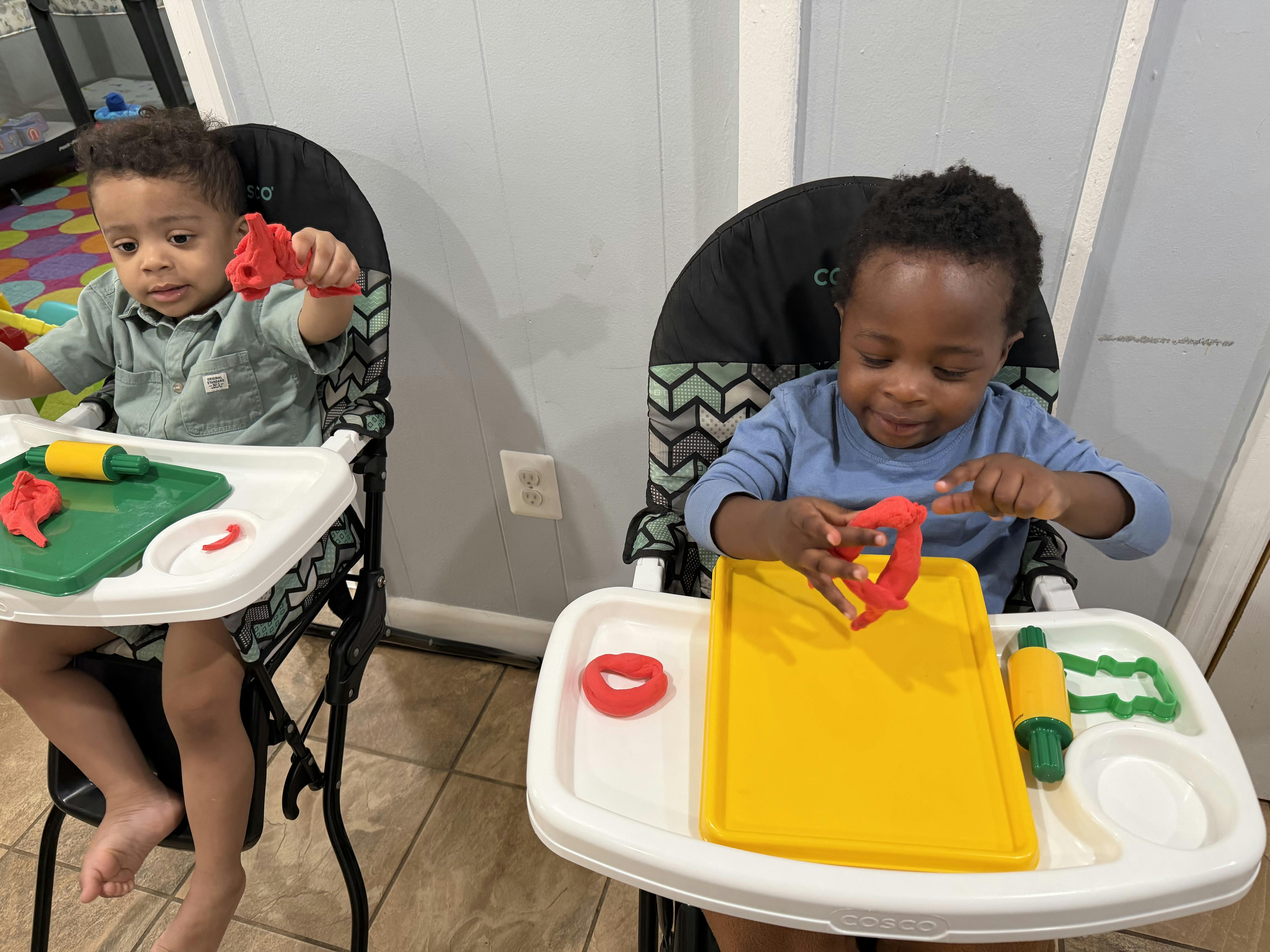 Two children sitting in high chairs playing with red modeling clay