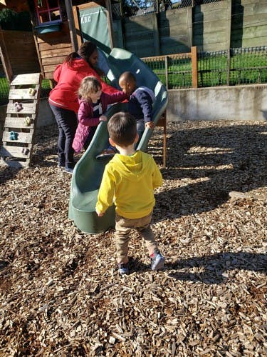 Person helping two children on a playground slide as another child watches