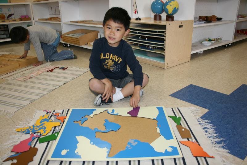 Child sitting on floor with a map puzzle, another child in background