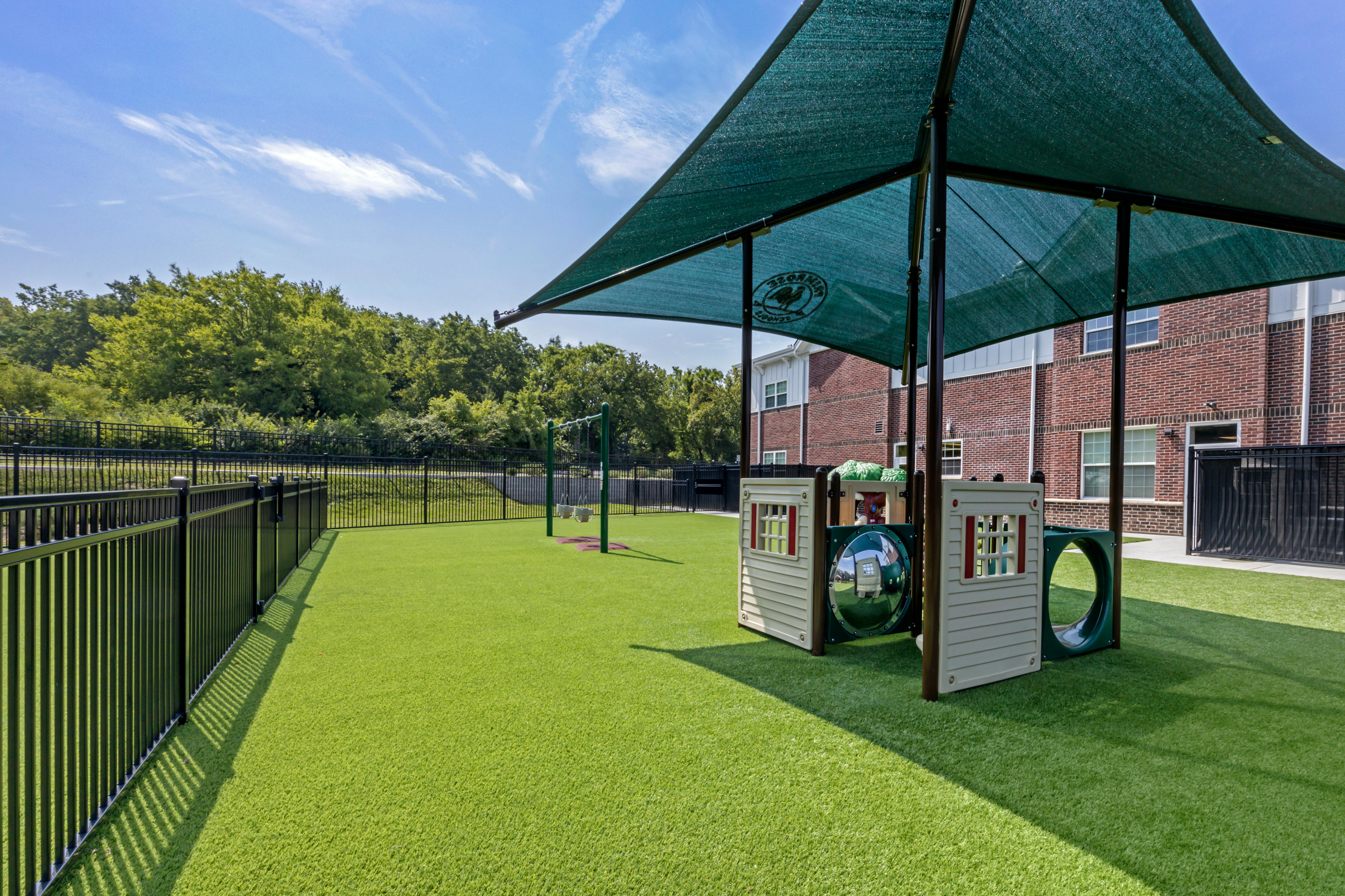 Playground with green turf, swing set, and small playhouse structure with a shade canopy