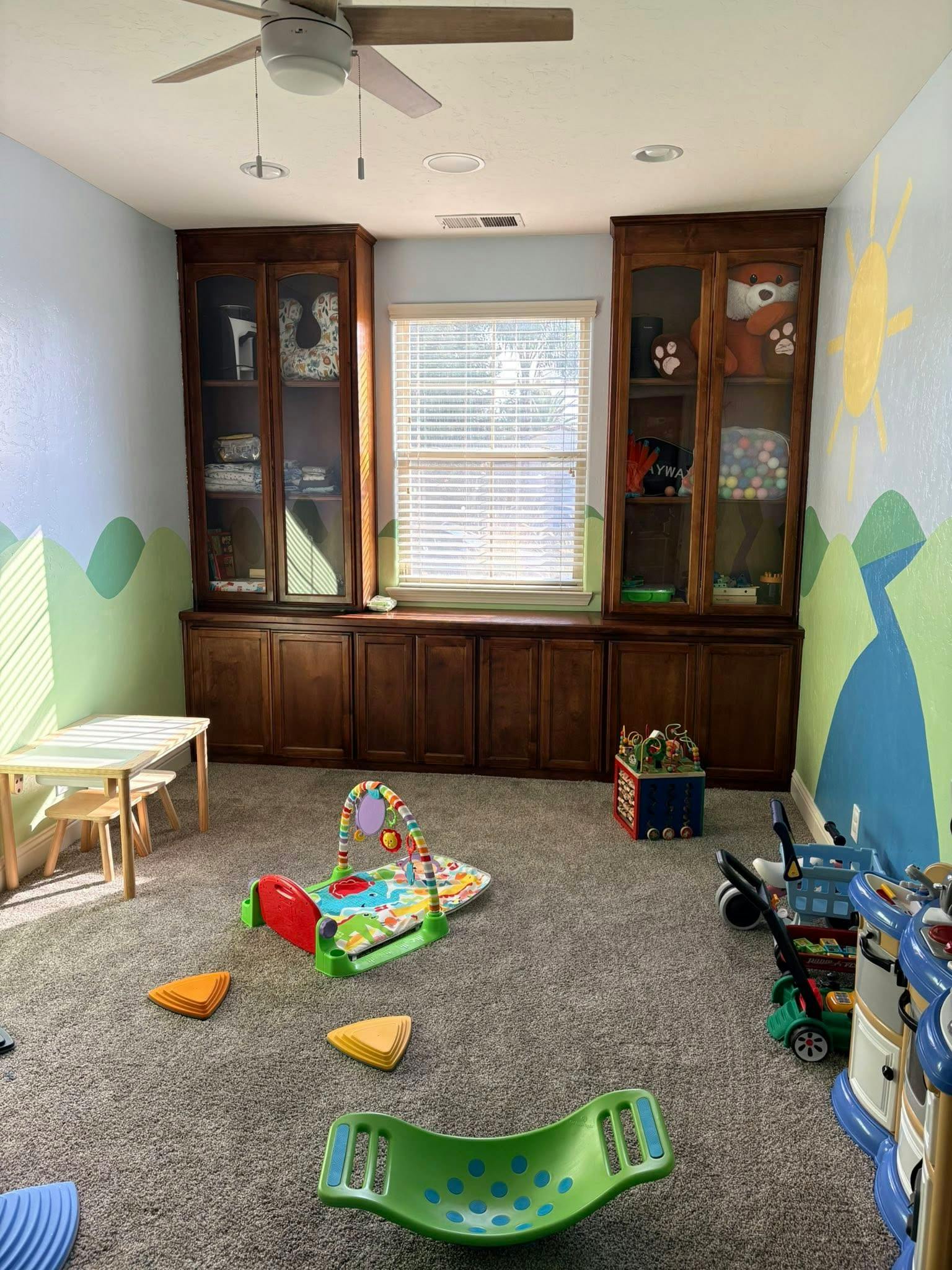 A playroom with built-in wooden cabinets, a window, and various toys on the carpet