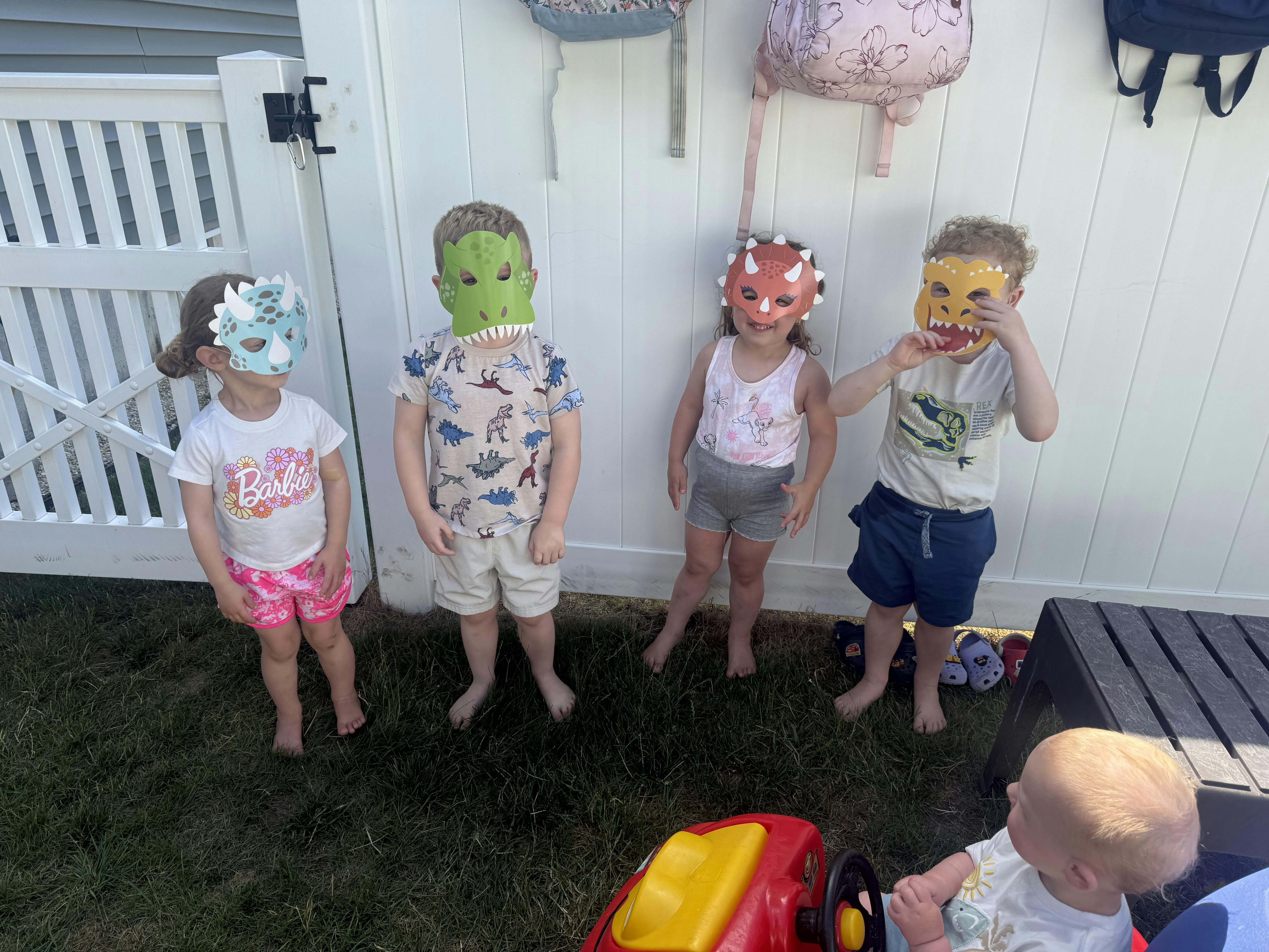 Four children wearing dinosaur masks standing in a row in front of a white fence