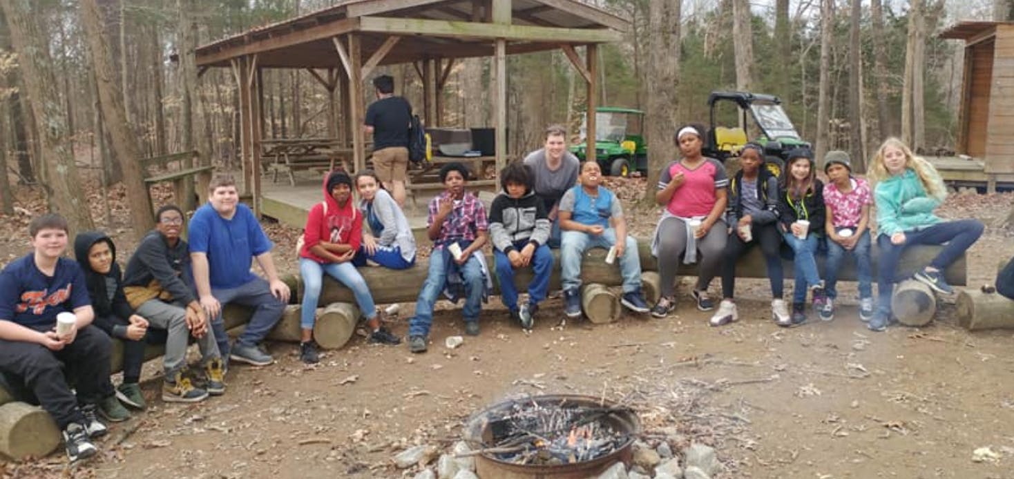 Group of teens and children sitting on logs around a campfire pit