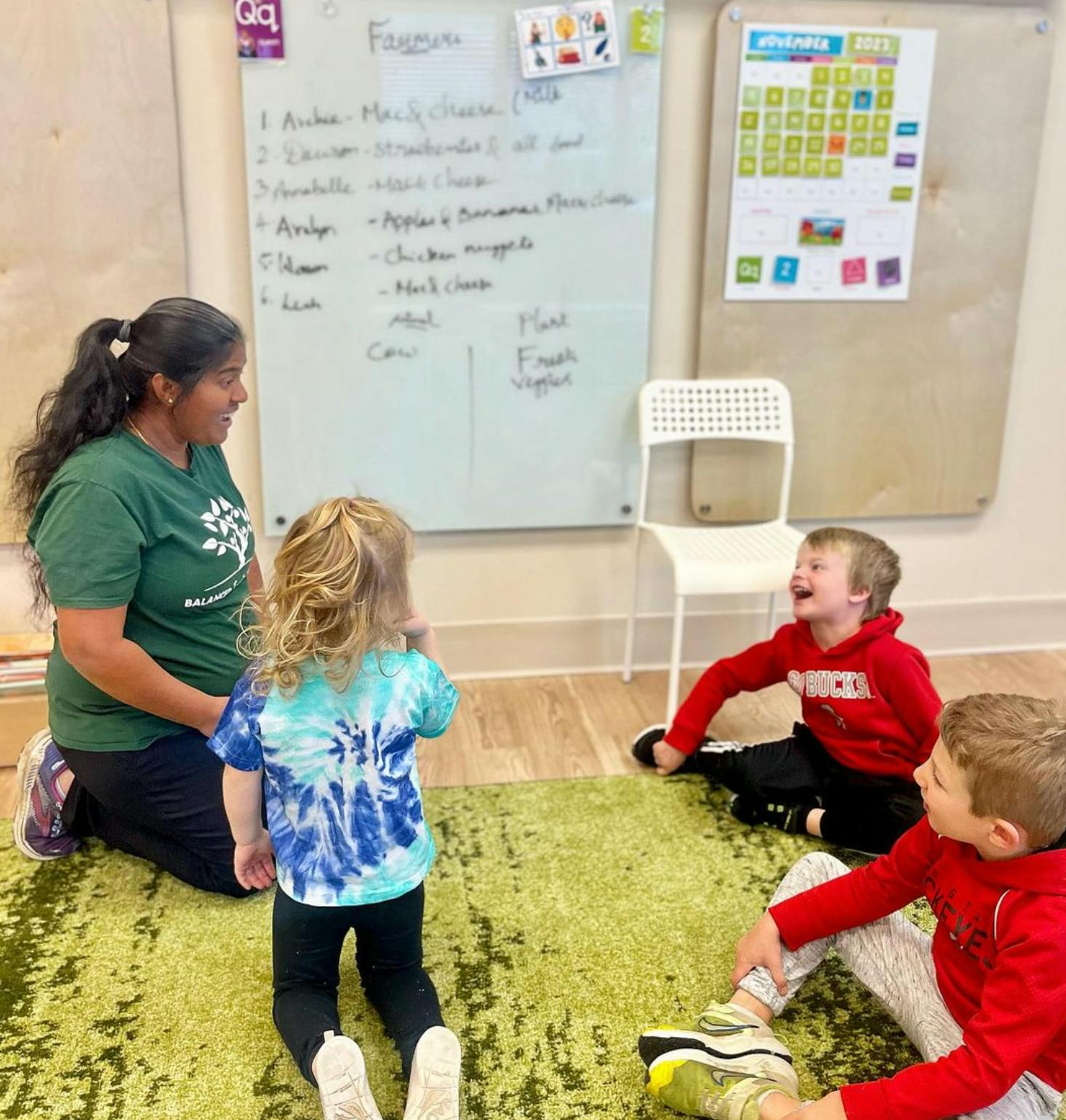 Person kneeling with three children sitting on a rug in a classroom