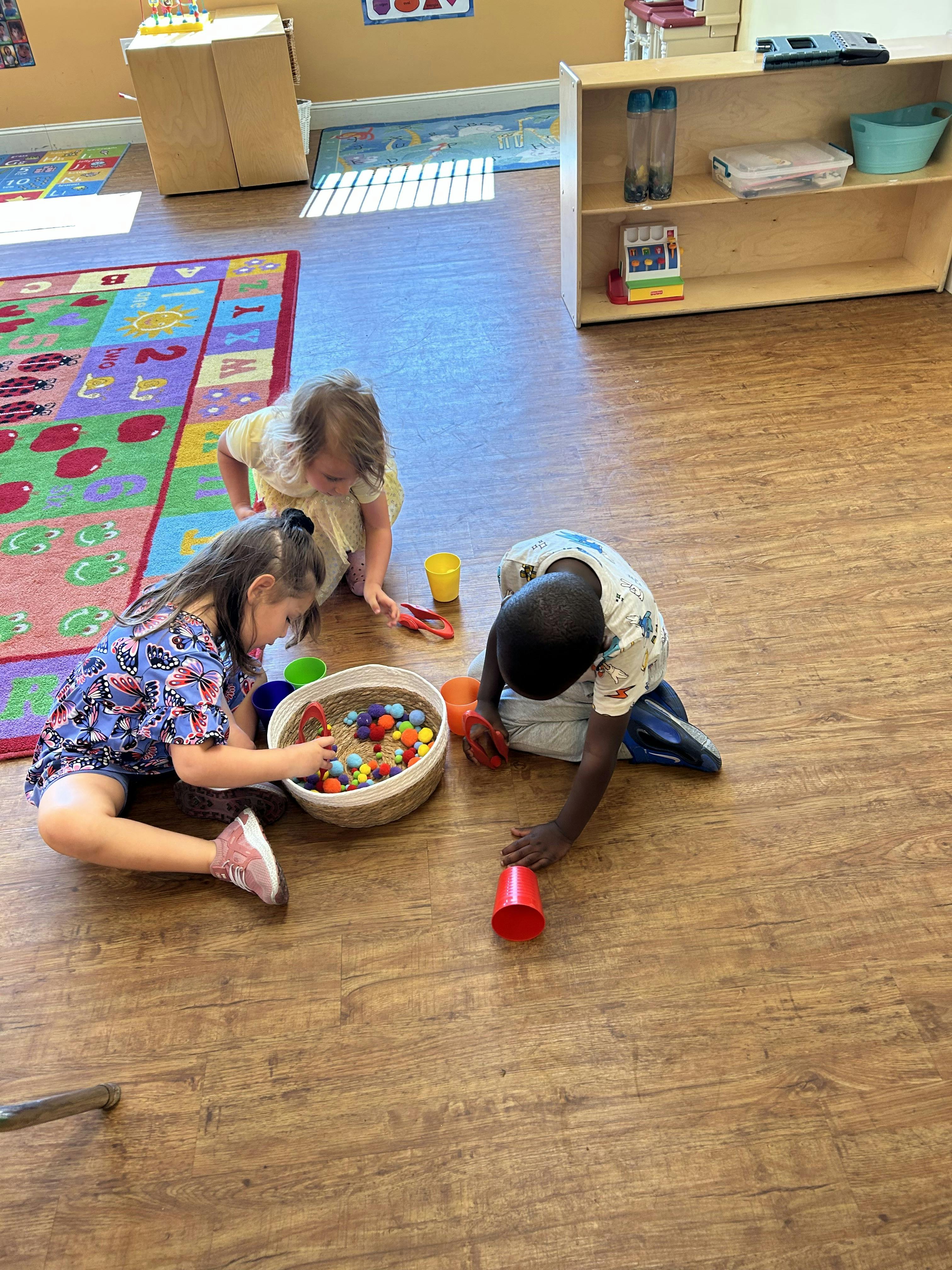 Three children playing with colorful small pom-poms and cups on the floor