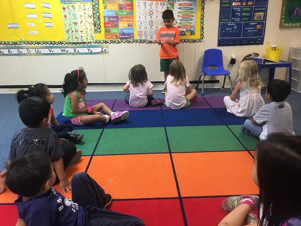 Child standing in front of other children sitting on a colorful mat in a classroom