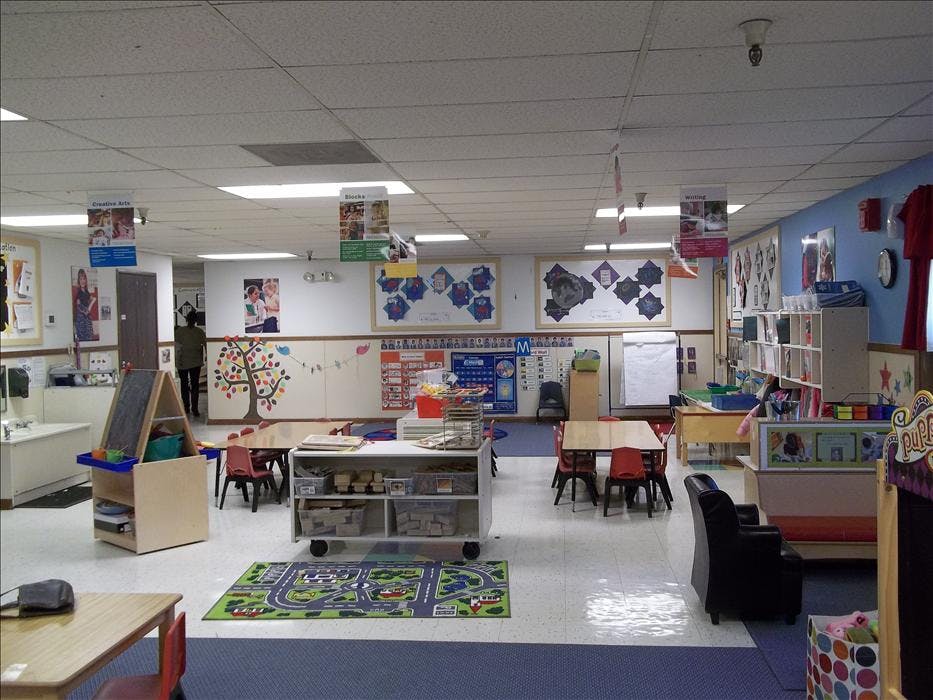 Classroom with small tables and chairs, a play rug, and activity displays on walls