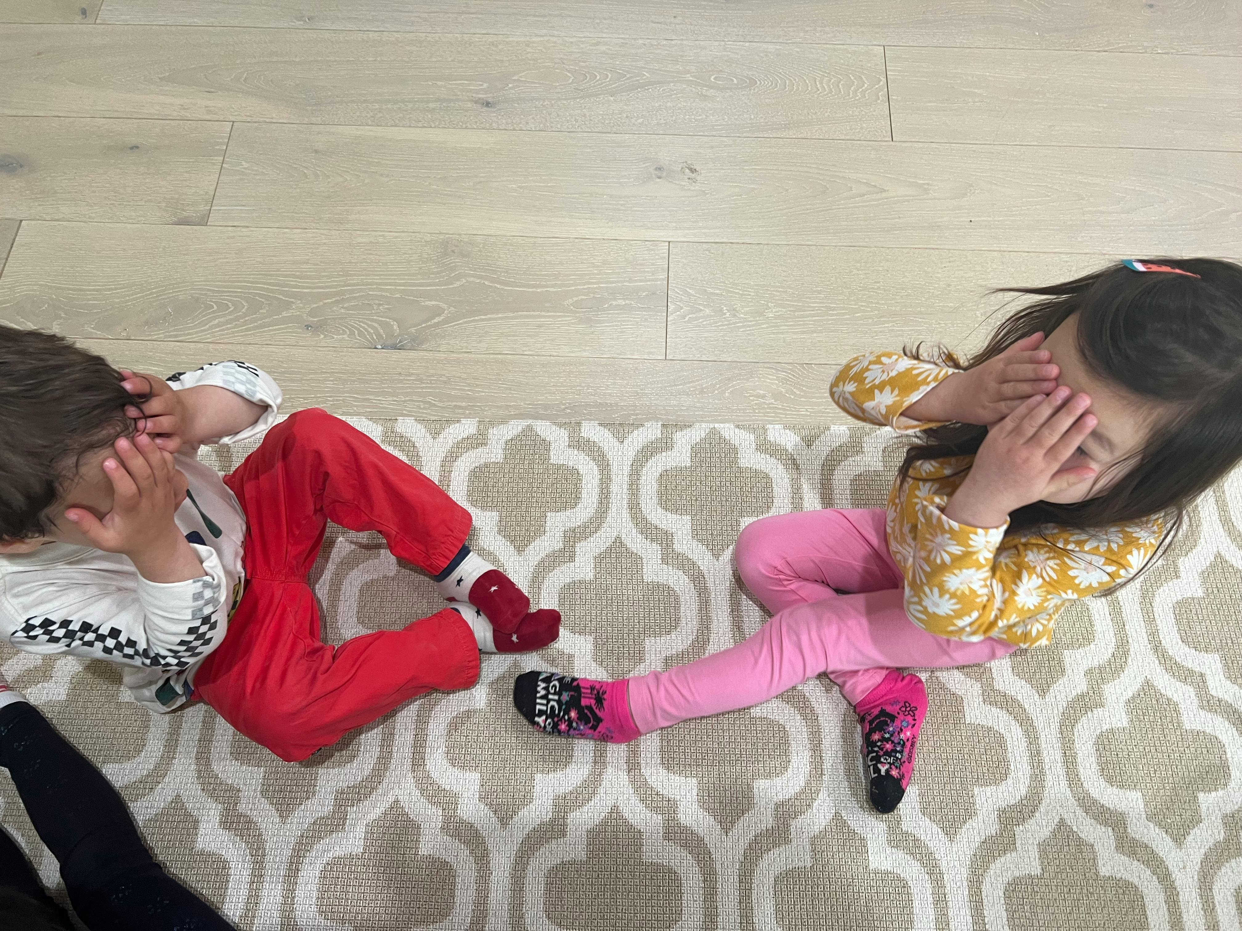 Two children lying on a rug with hands covering their faces