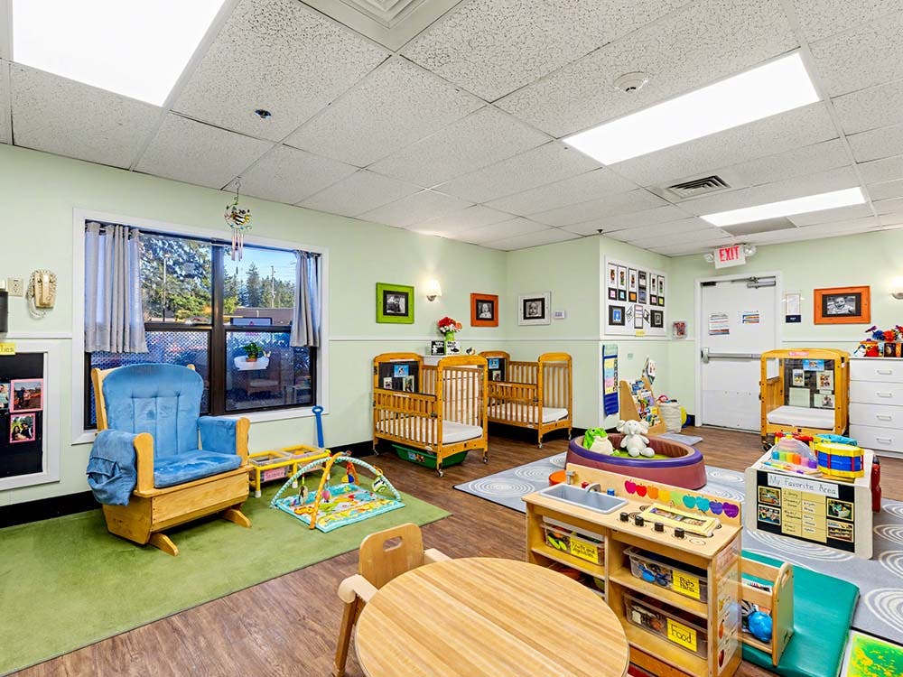 A brightly lit nursery room with multiple cribs, a rocking chair, and play areas
