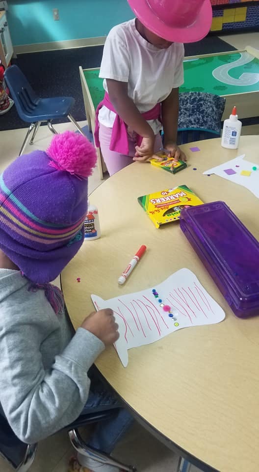Two children at a table doing crafts with crafts, one drawing and one handling a box