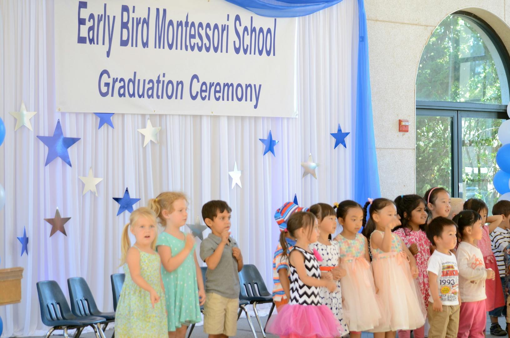 Children standing in a line under a banner that reads "Early Bird Montessori School Graduation Ceremony"