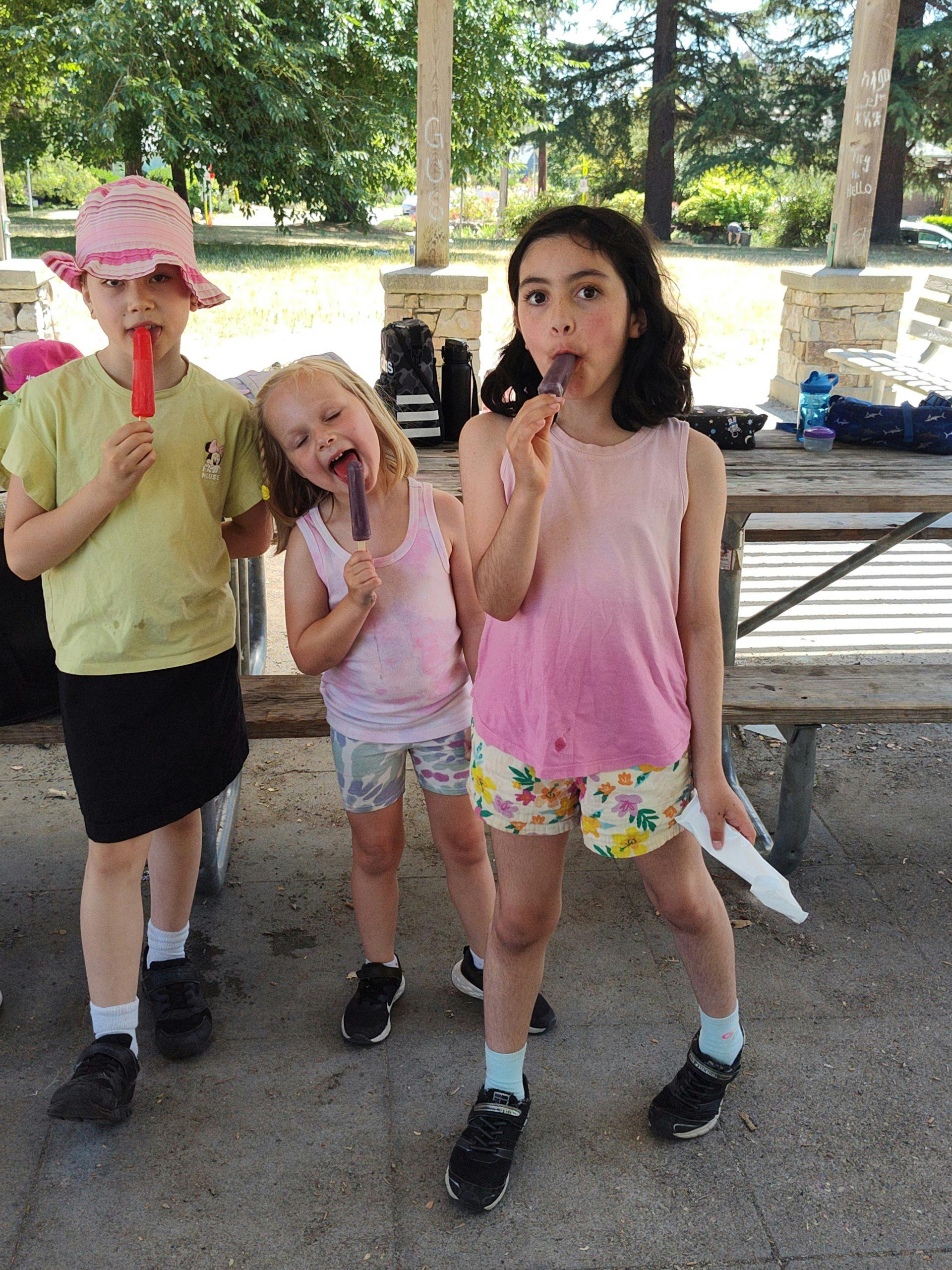 Three children eating popsicles under a shelter