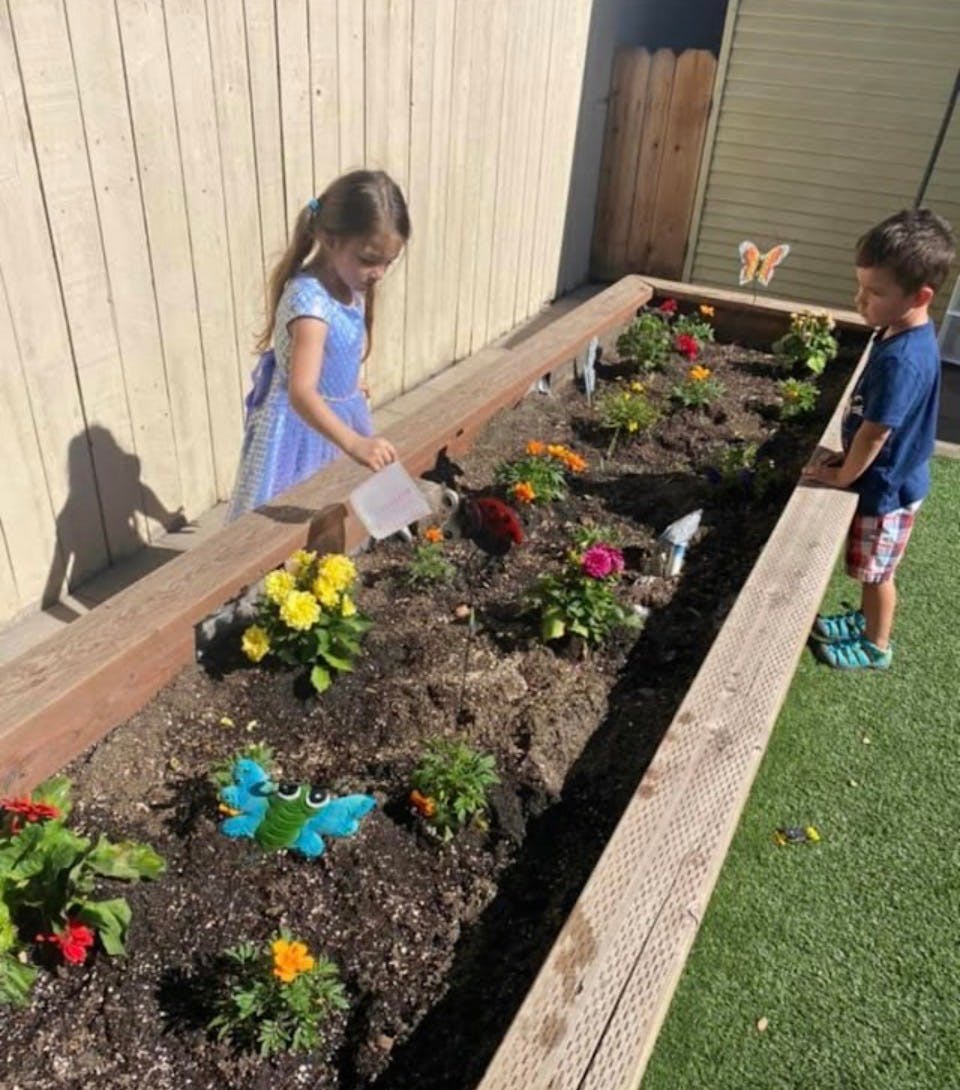 Child watering plants in a raised garden bed with another child nearby
