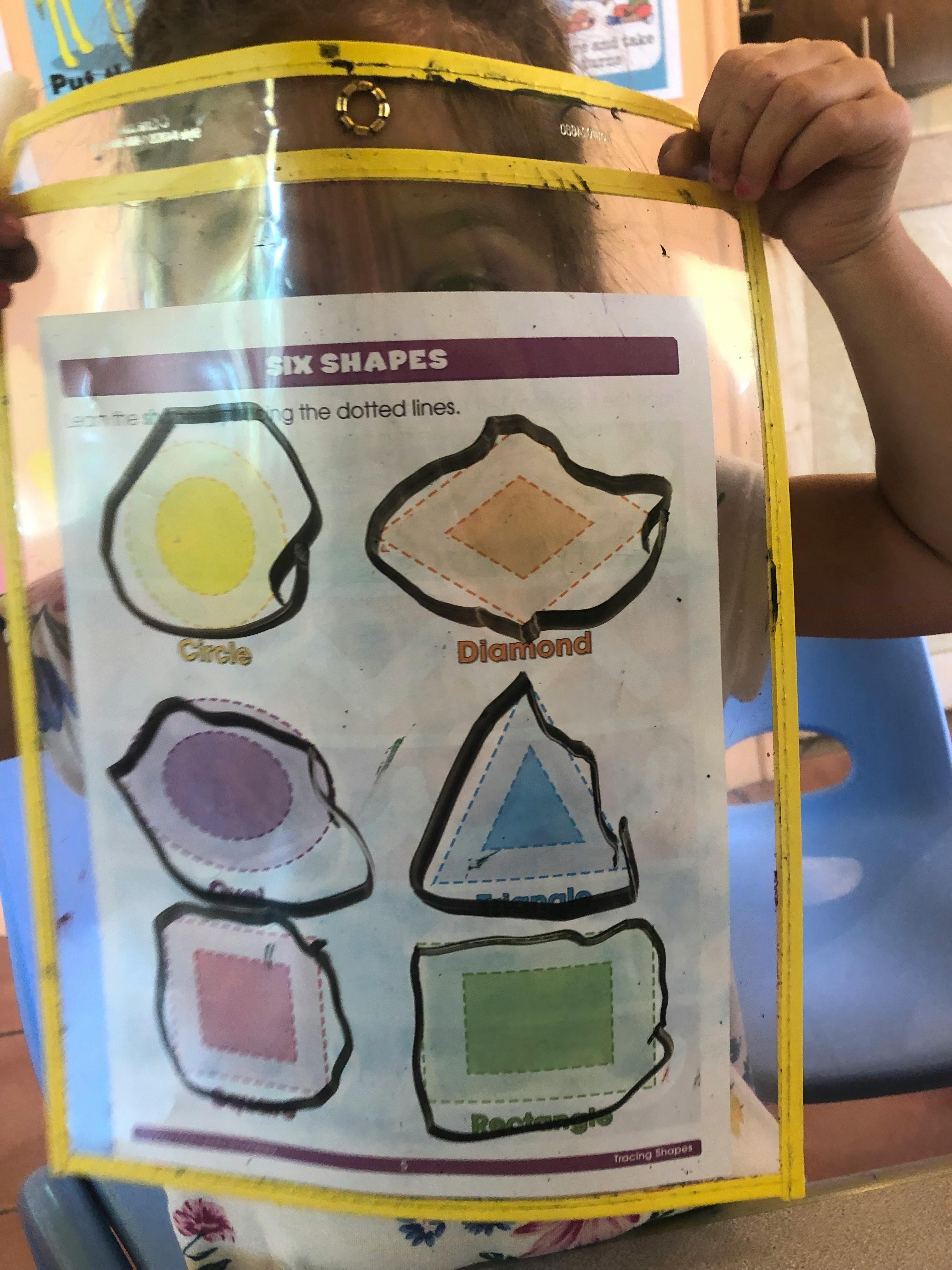 Child holding up a clear plastic sleeve with a "Six Shapes" worksheet inside