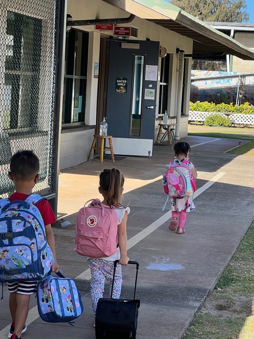 Three children with backpacks walking on a sidewalk toward a building