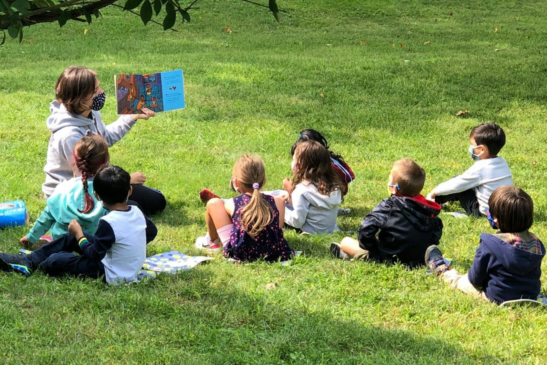 Person reading a book to a group of children sitting on grass