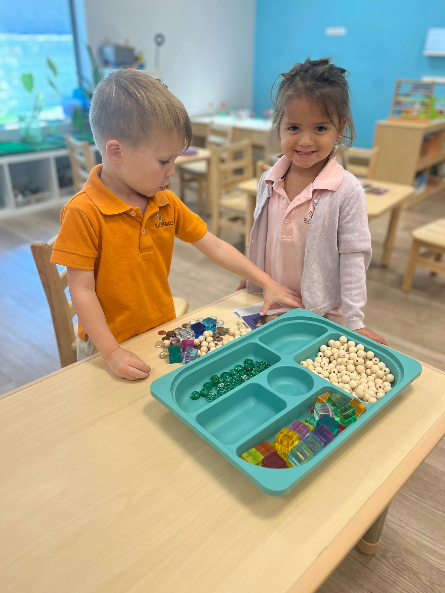Two children standing at a table with a tray of beads and craft supplies