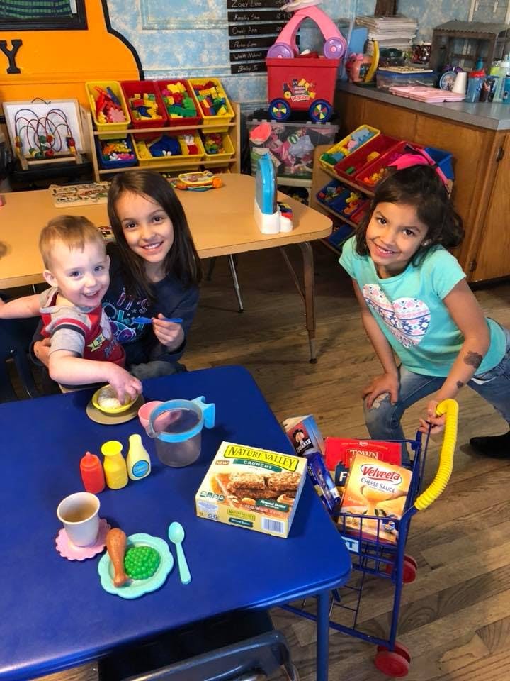 Two children and a teen playing with toy food and a shopping cart in a classroom