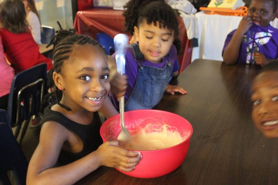 Child stirring batter in a red bowl with other children nearby