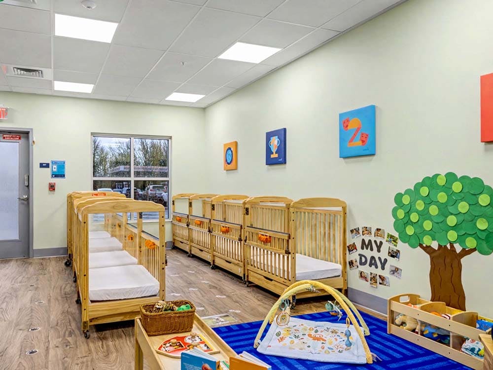 Classroom with multiple wooden cribs, an activity mat, and a tree decoration on the wall