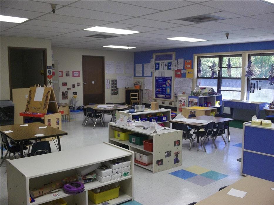 Empty classroom with small tables and chairs, shelves with toys and supplies
