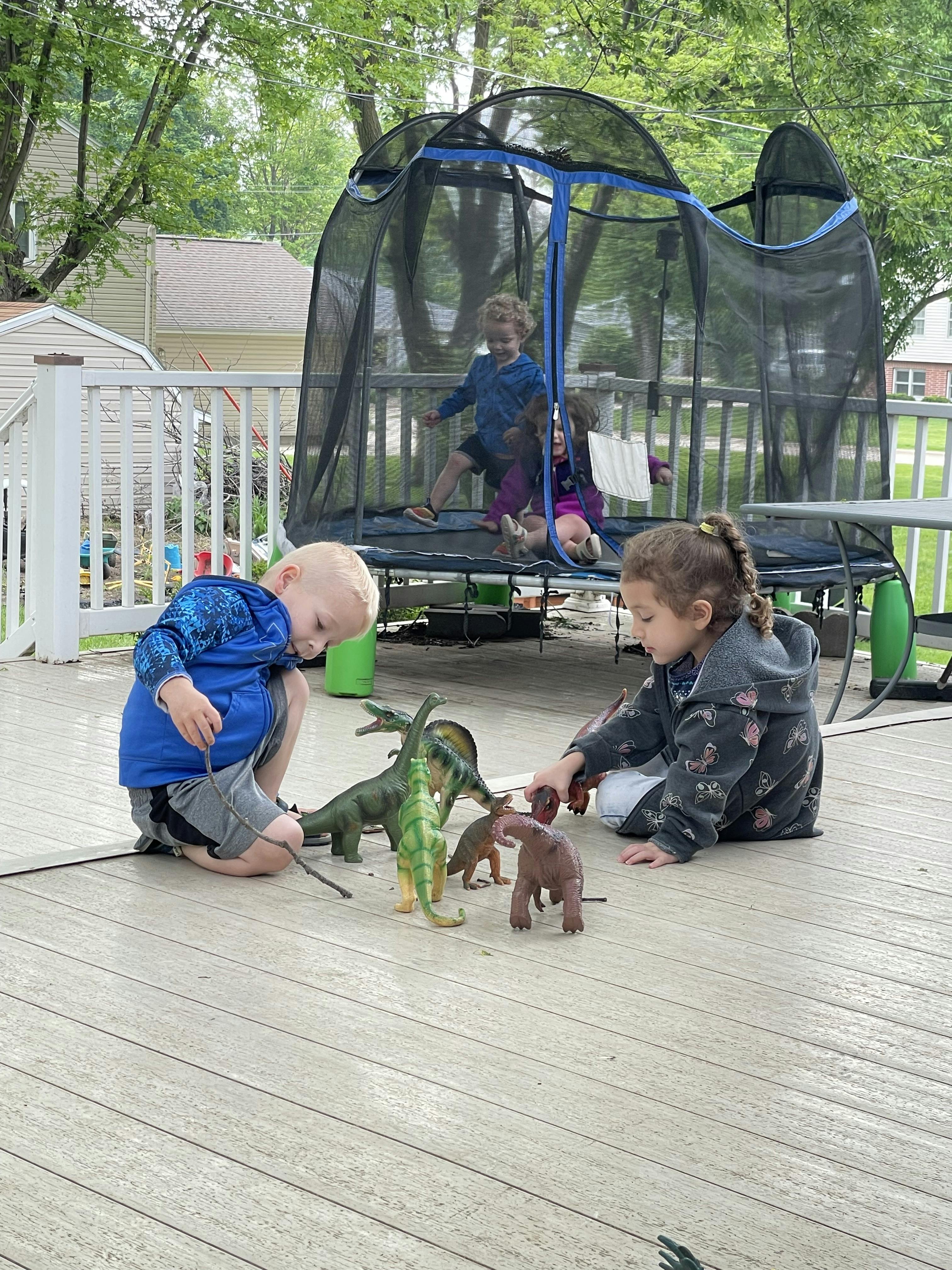 Children playing with dinosaur toys on a deck, with others inside a trampoline