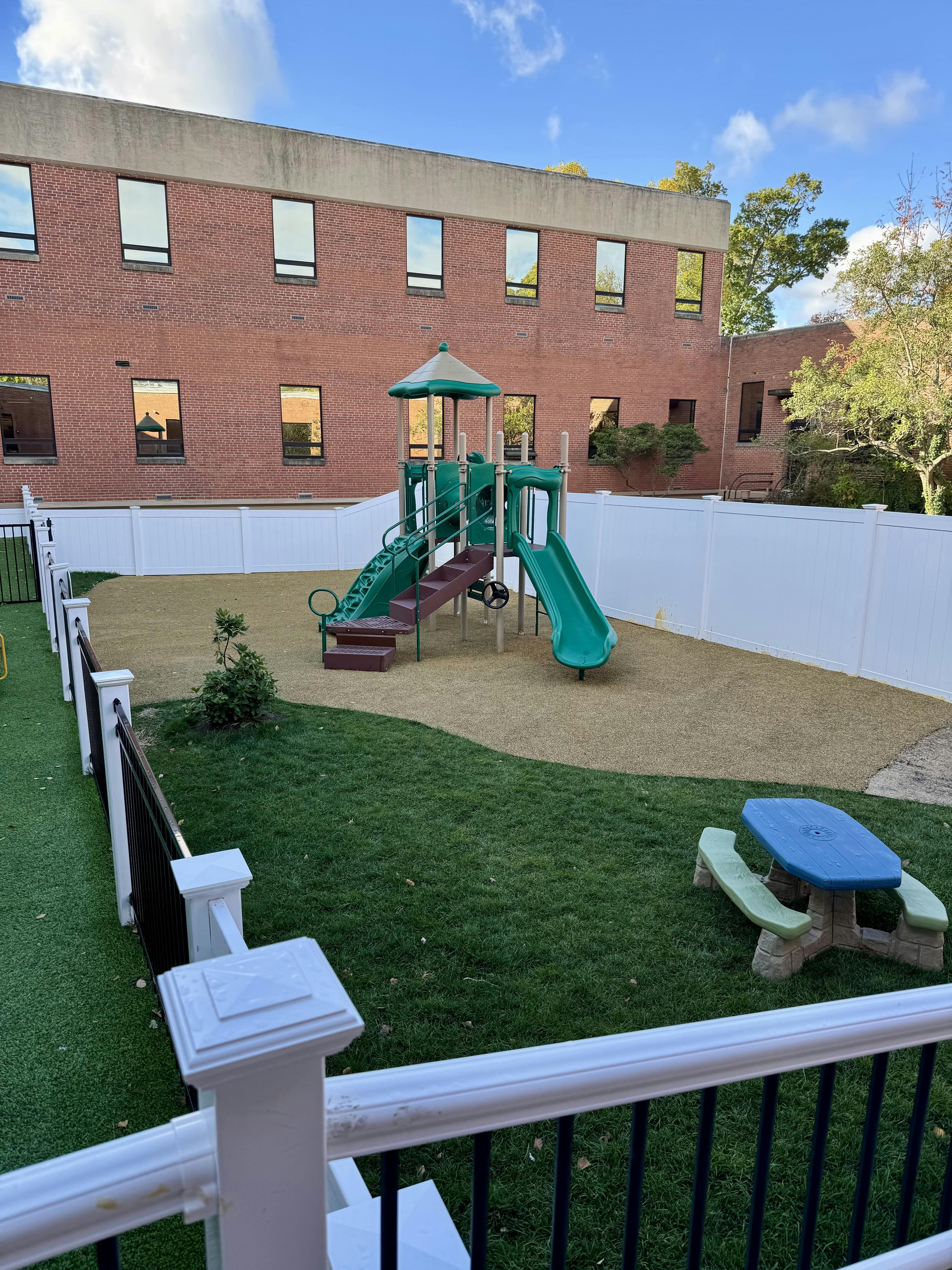 A playground with a green and brown playset, a small blue and green picnic table, and a brick building in the background