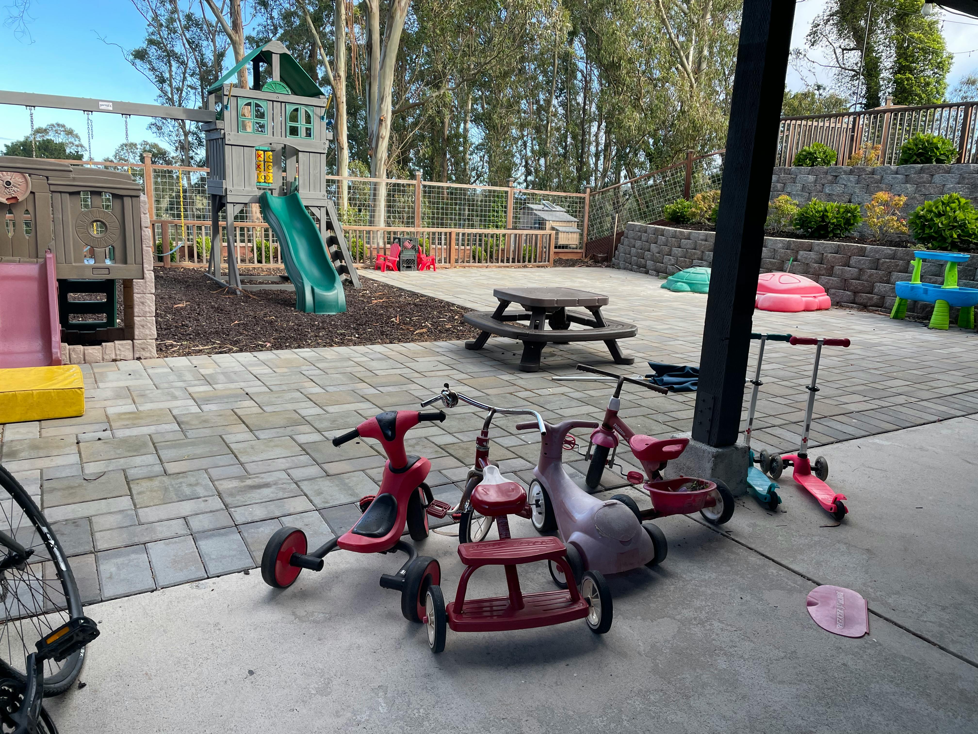 Tricycles and scooters on a paved area next to a playground