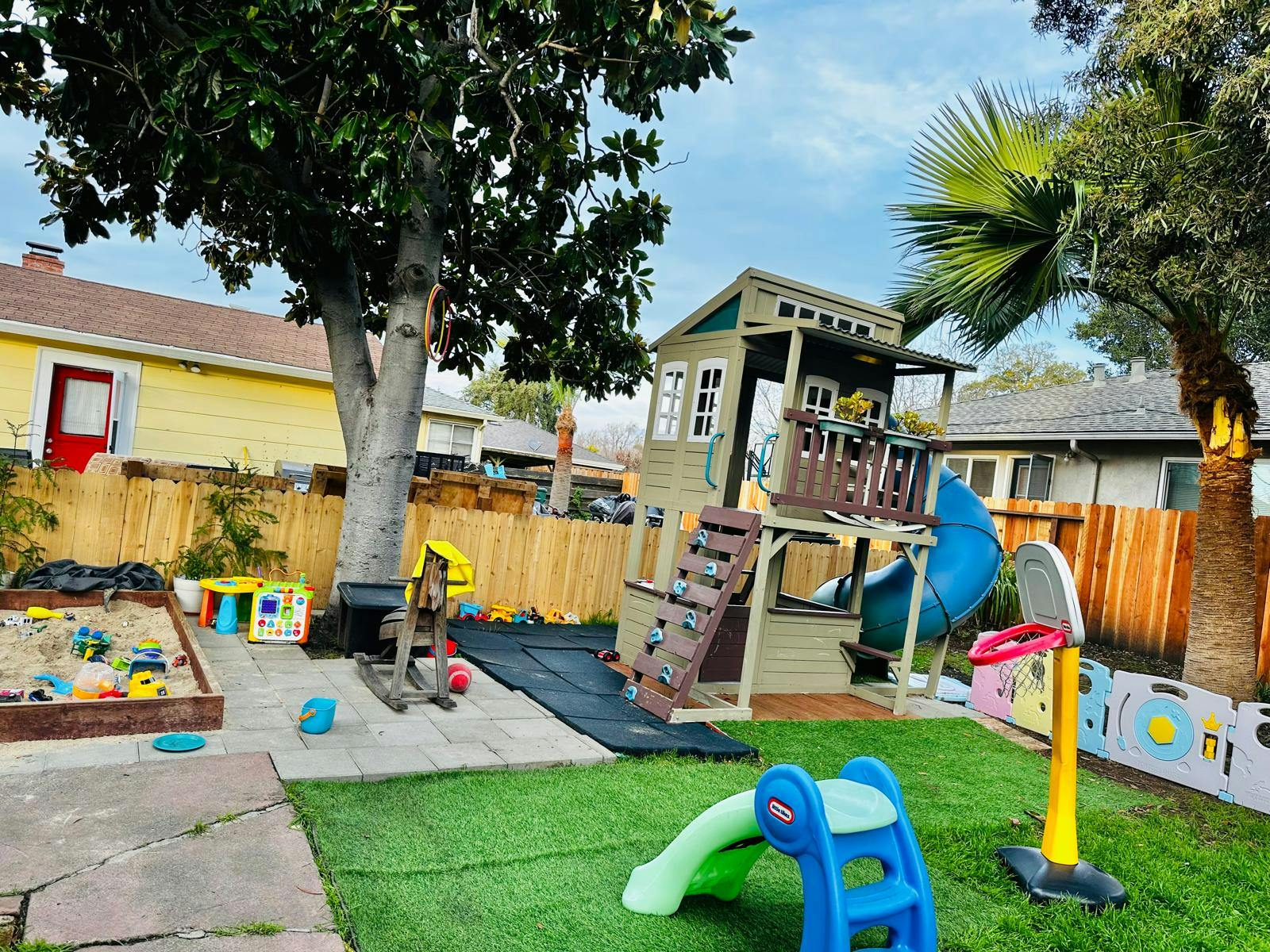Outdoor playground with a wooden playhouse, slide, sandbox, and artificial turf
