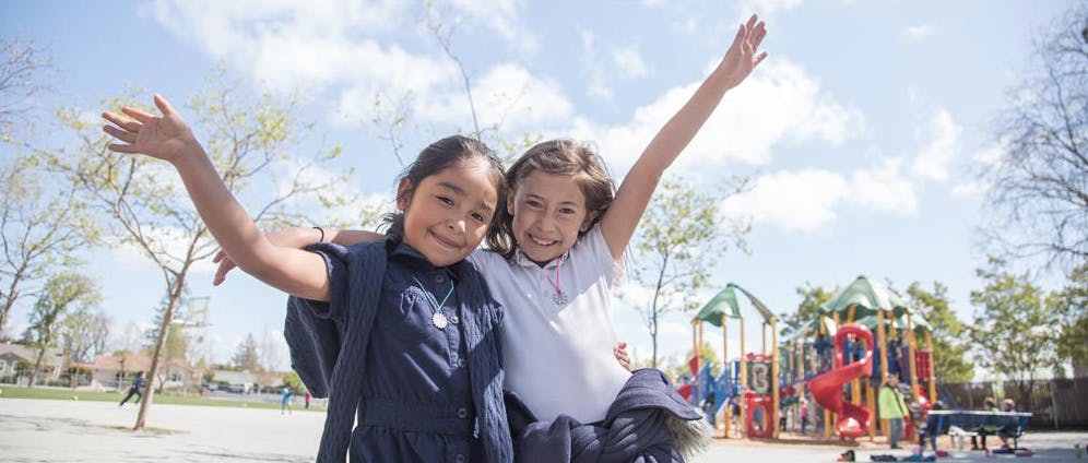 Two children with arms raised in front of a playground