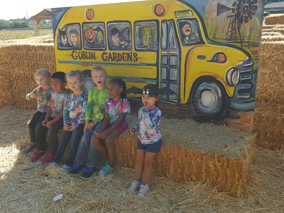 Children sitting on hay bales in front of a school bus cutout with "Goblin Gardens" on it