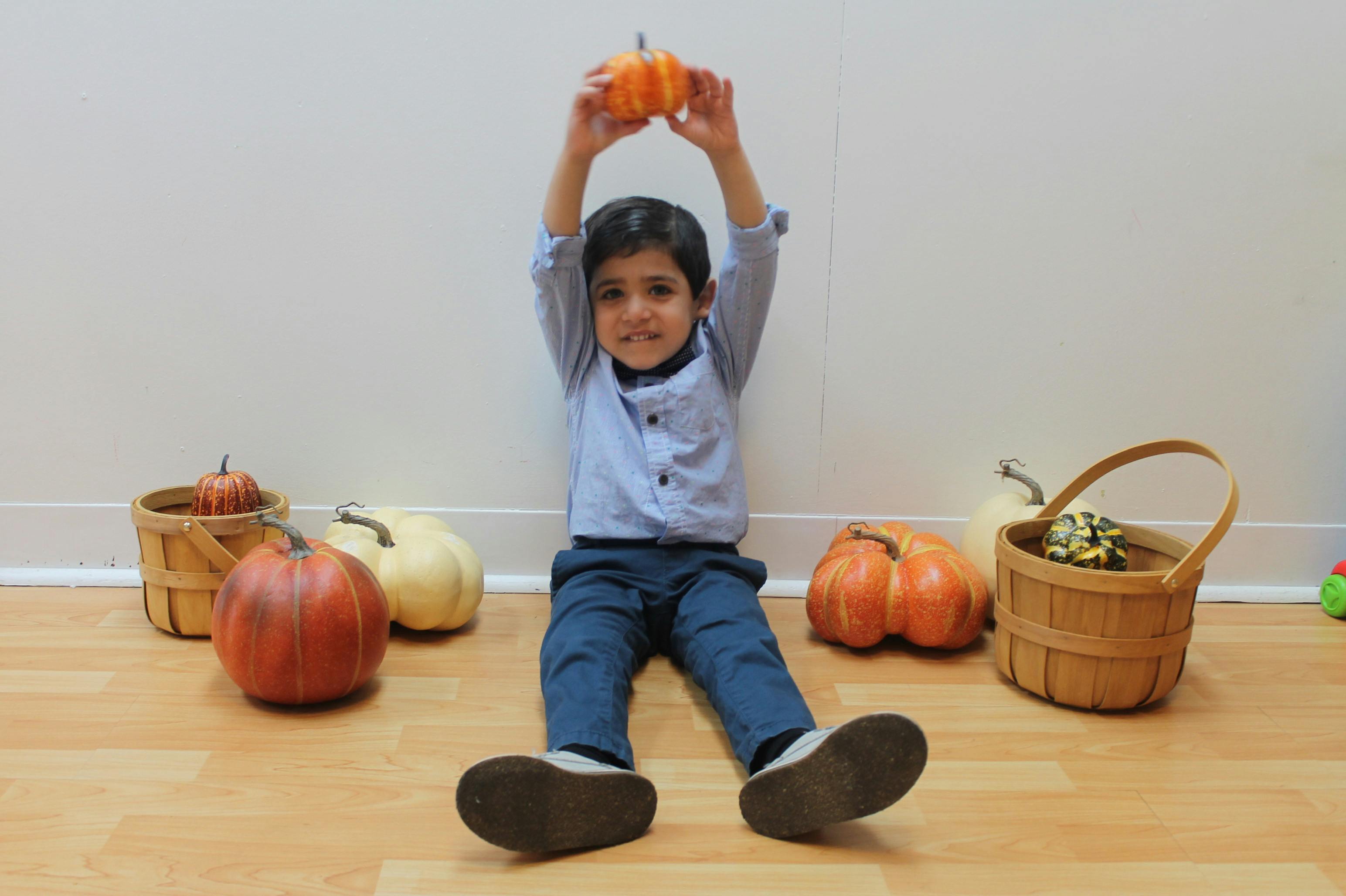 Child sitting on floor holding a small pumpkin above their head