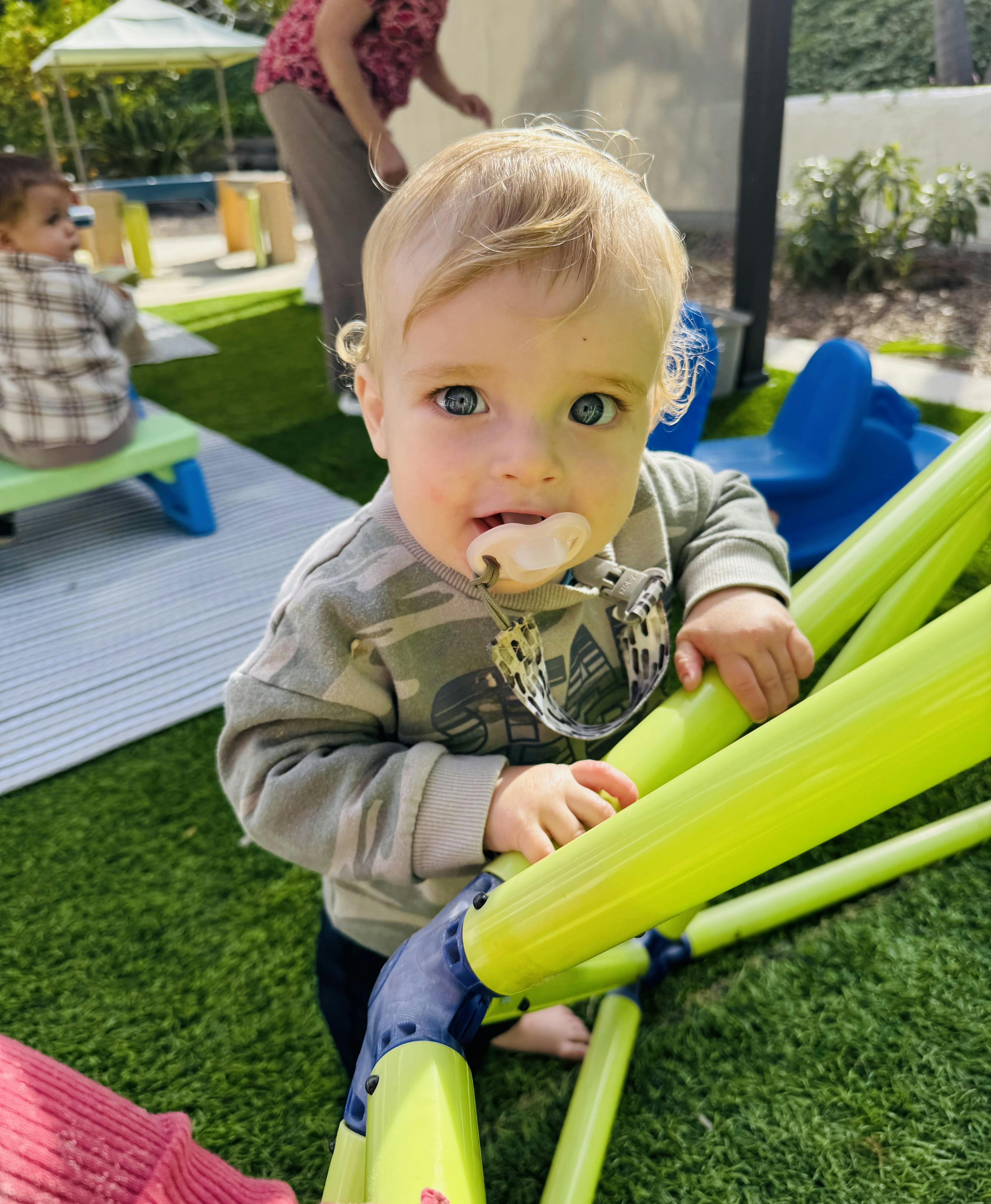 Child with a pacifier in mouth, holding onto a green play structure
