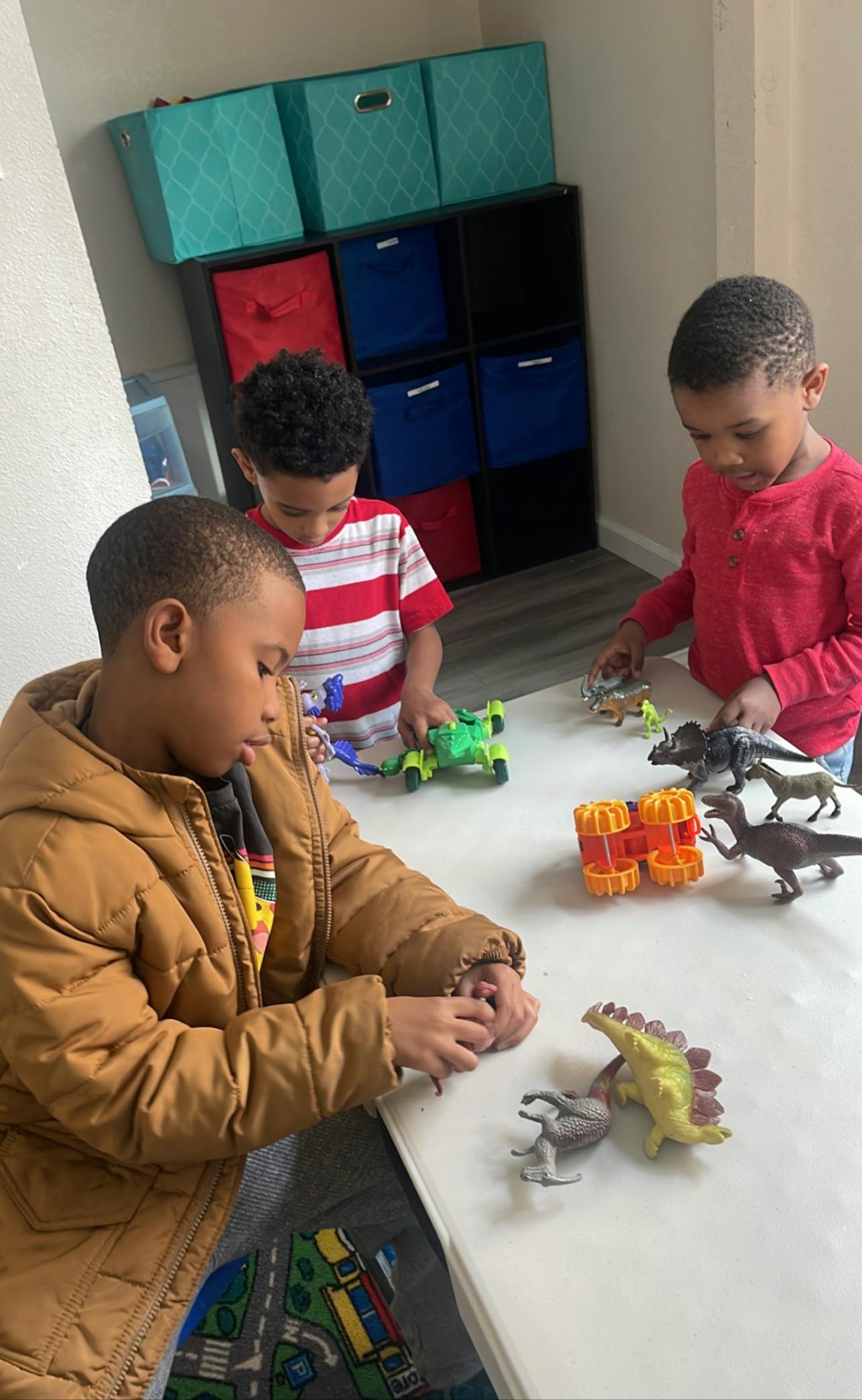 Three children playing with toy dinosaurs and vehicles on a table