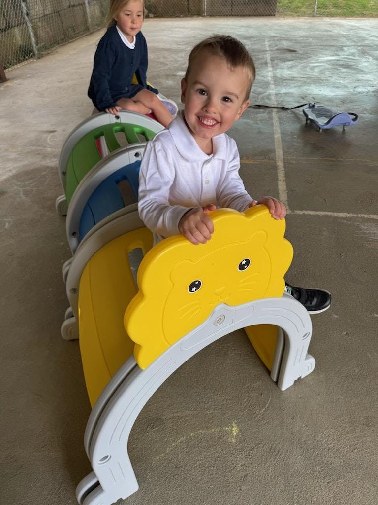 Child smiling while sitting on a toy lion, with another child in the background