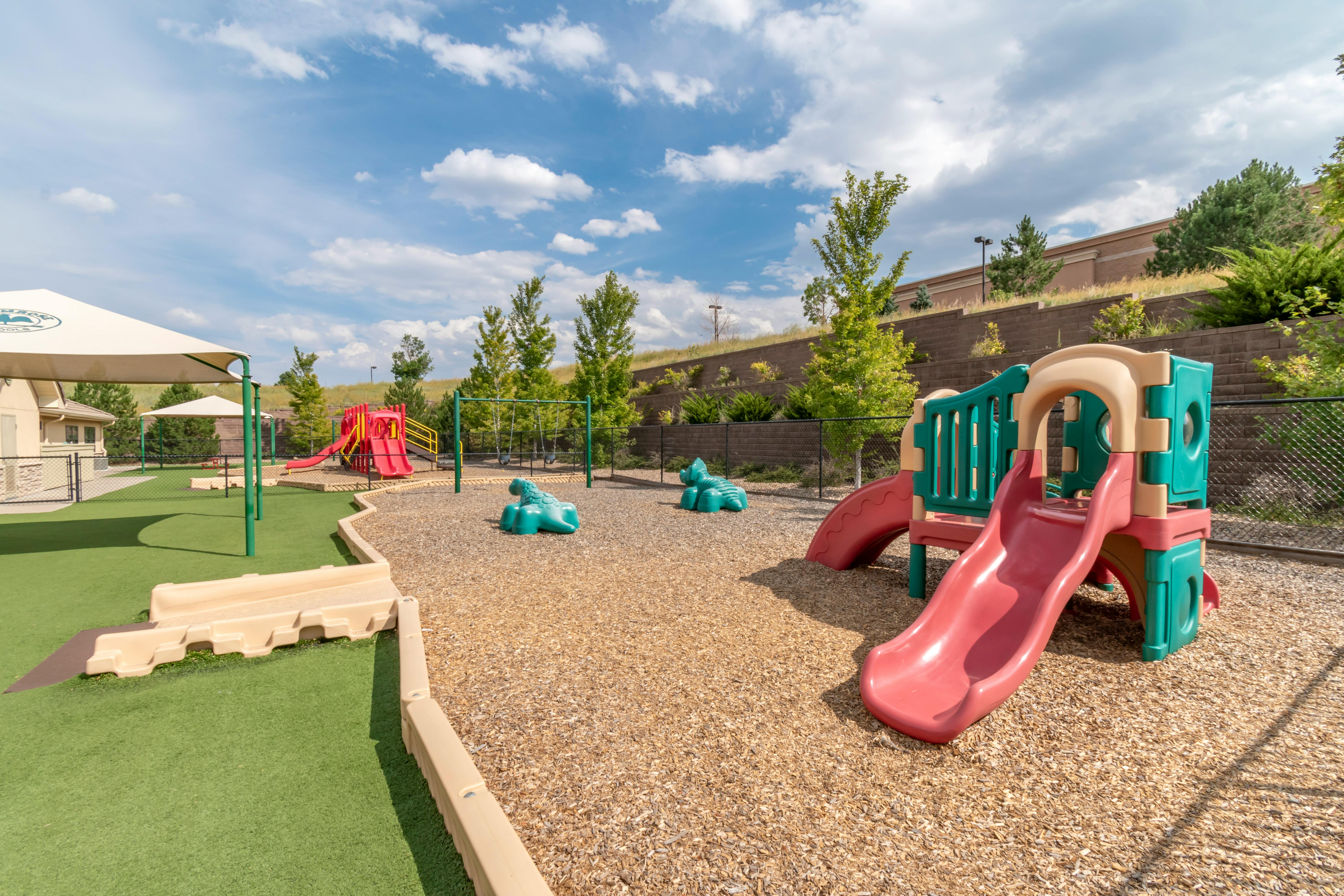 Outdoor playground with red slides, swings, and teal spring riders on wood chips