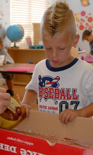 Child in a classroom looking down at a box, another person pouring liquid