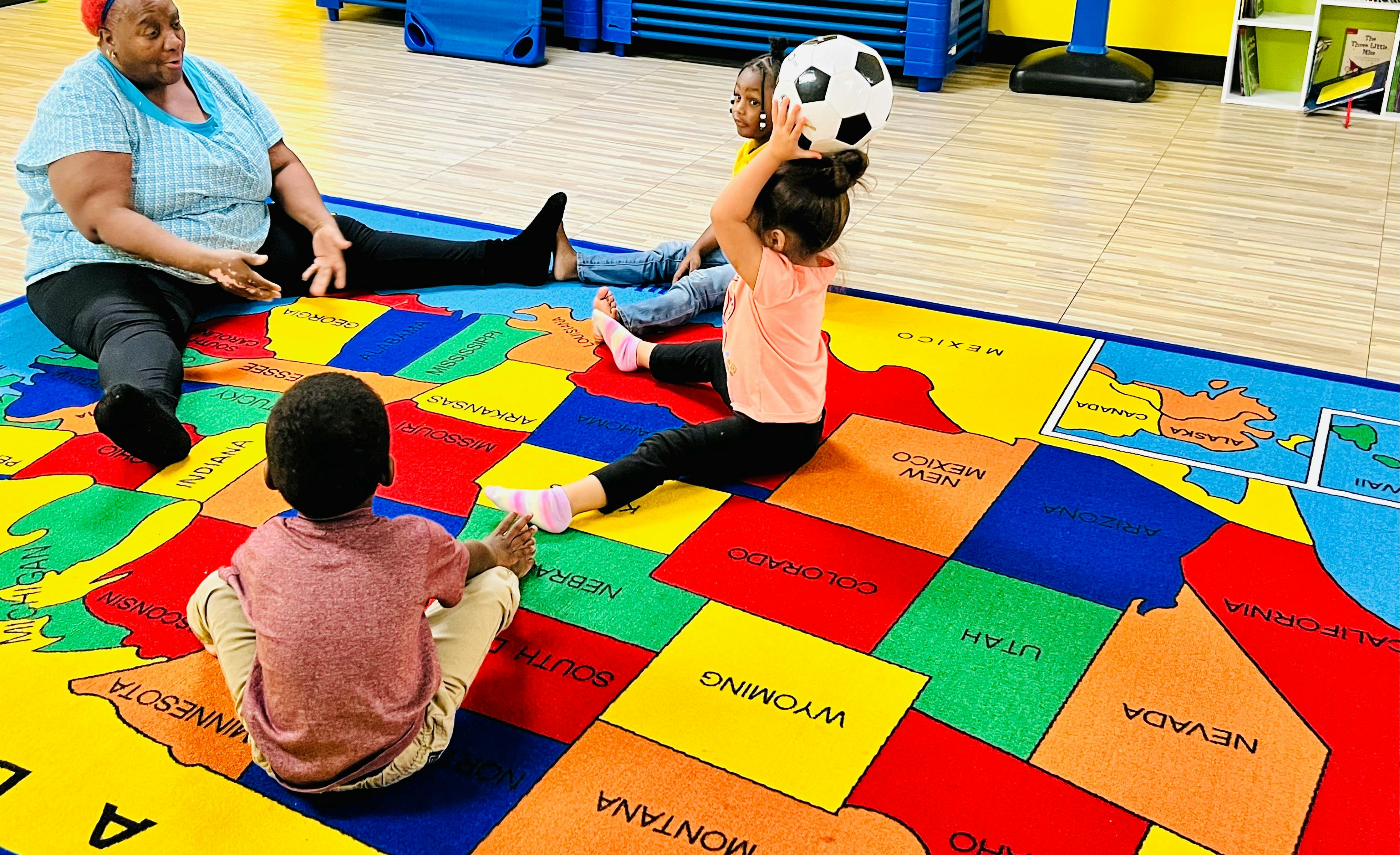 Adult and children sitting on a colorful map rug, child holding a soccer ball